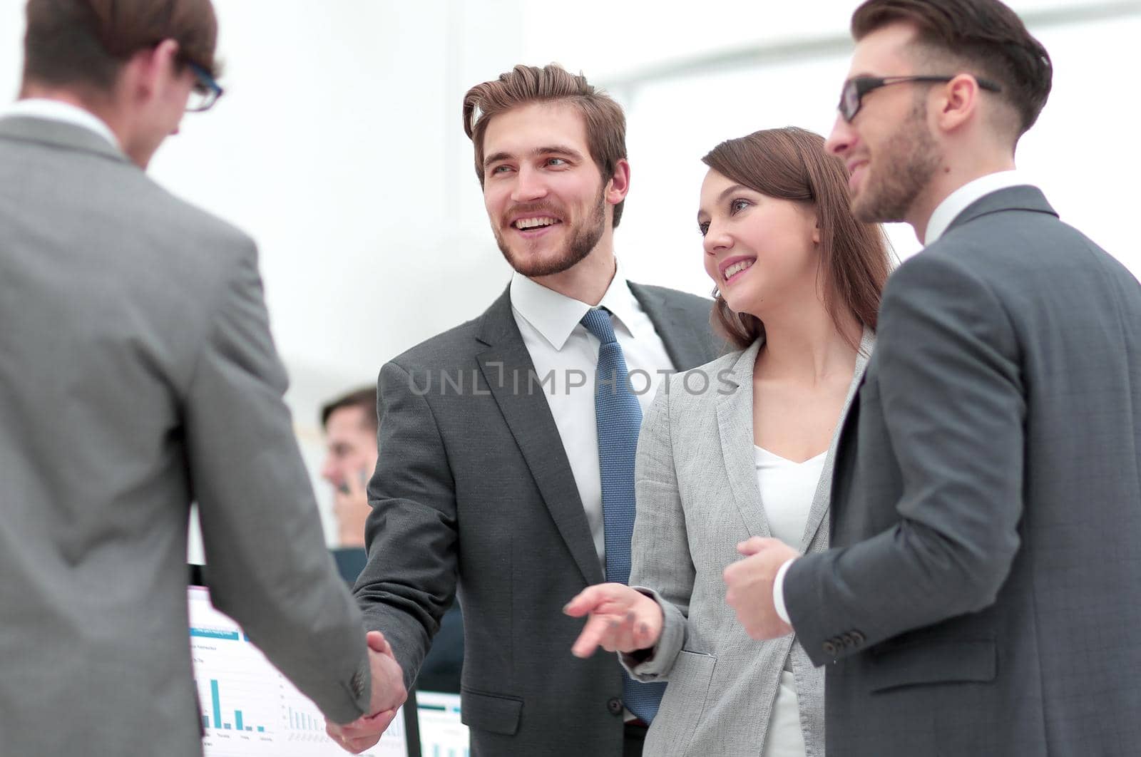 Welcome to team! Two men shaking hands and looking at each other with smile while their co workers sitting at the business meeting