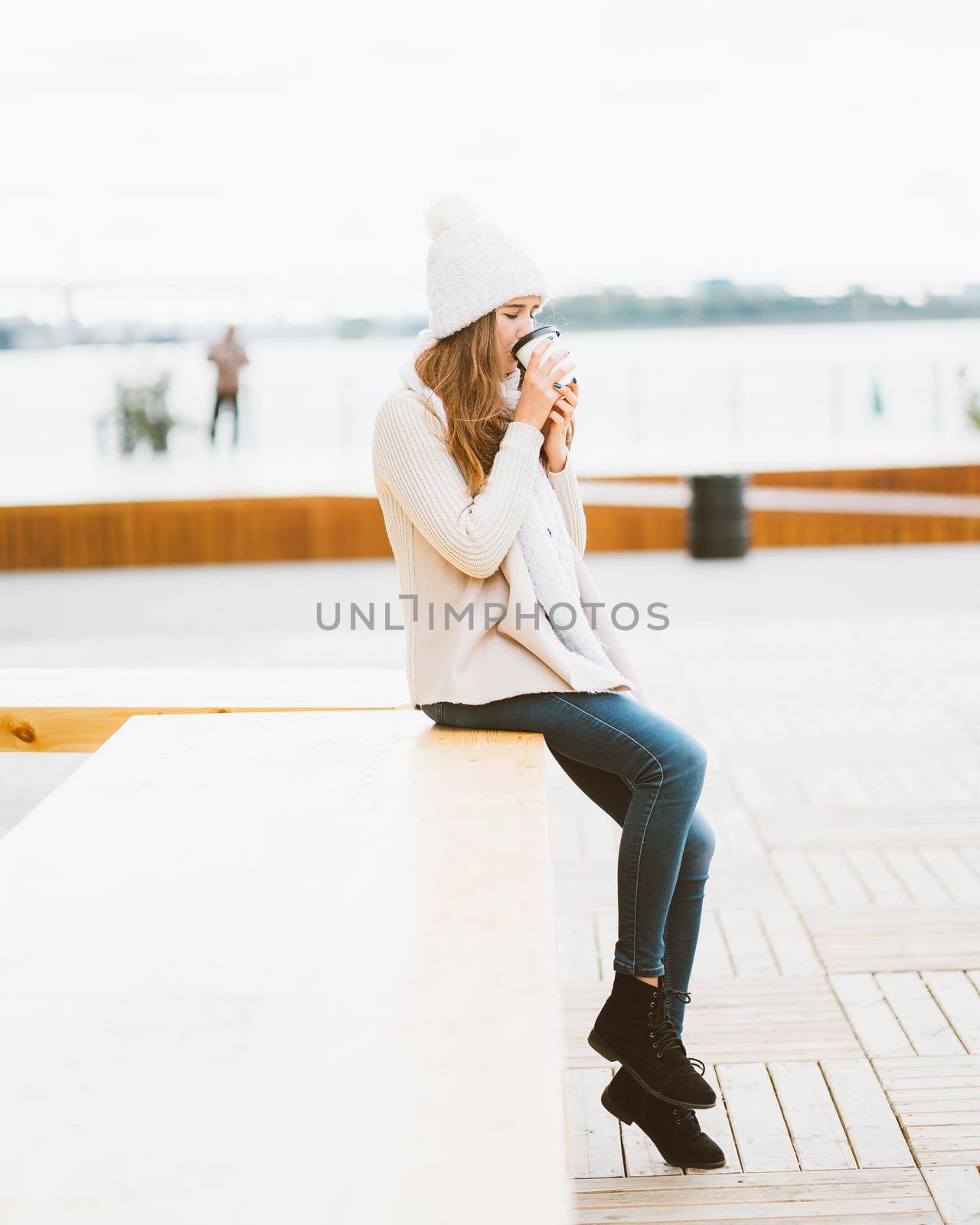 Beautiful young girl drinking coffee, tea from plastic mug in autumn, winter. A woman with long hair sitting on waterfront on Baltic sea in port and waiting for ferry, heated by hot drink, vertical by NataBene