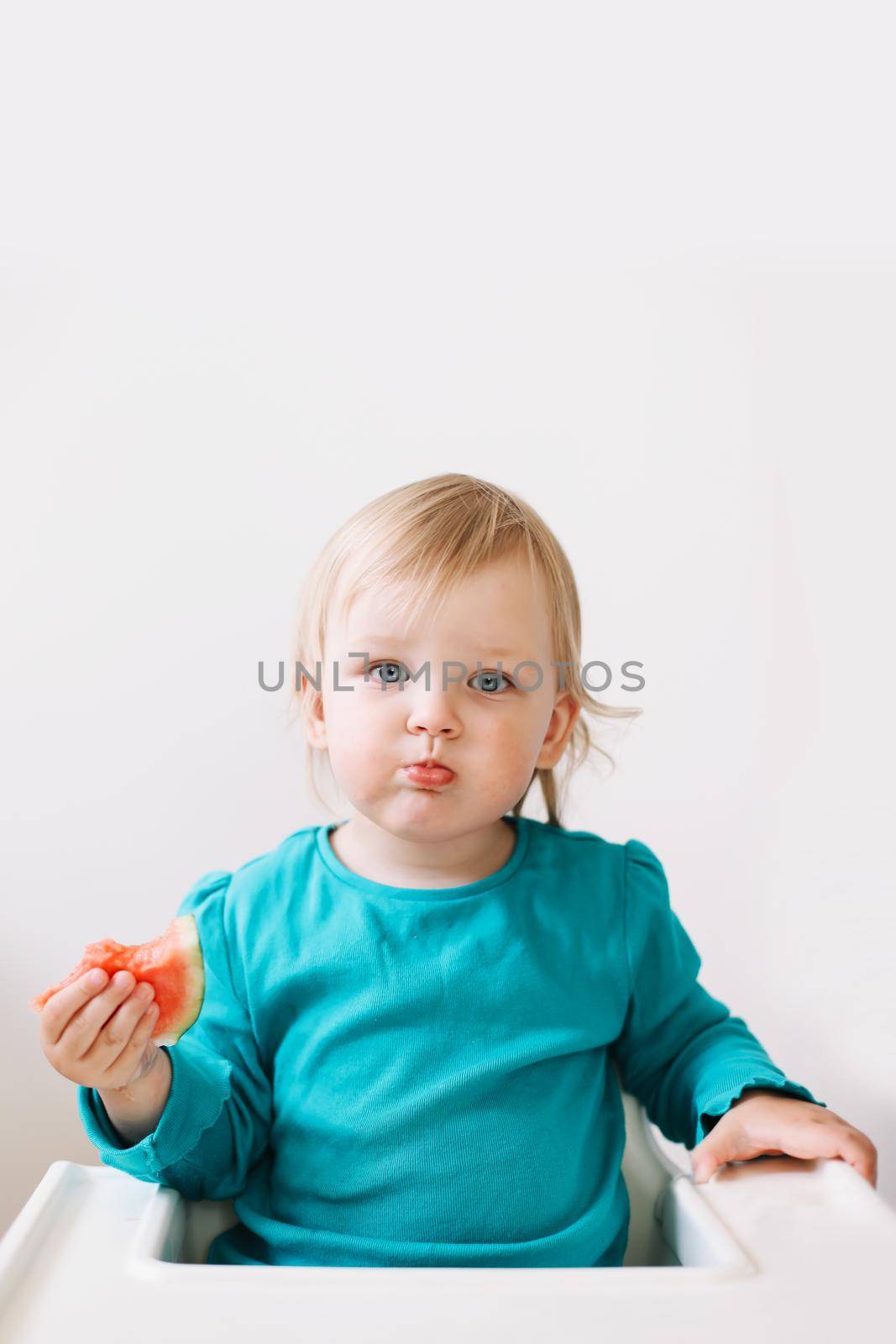 portrait of a funny little baby girl sitting in a high chair and eating watermelon.