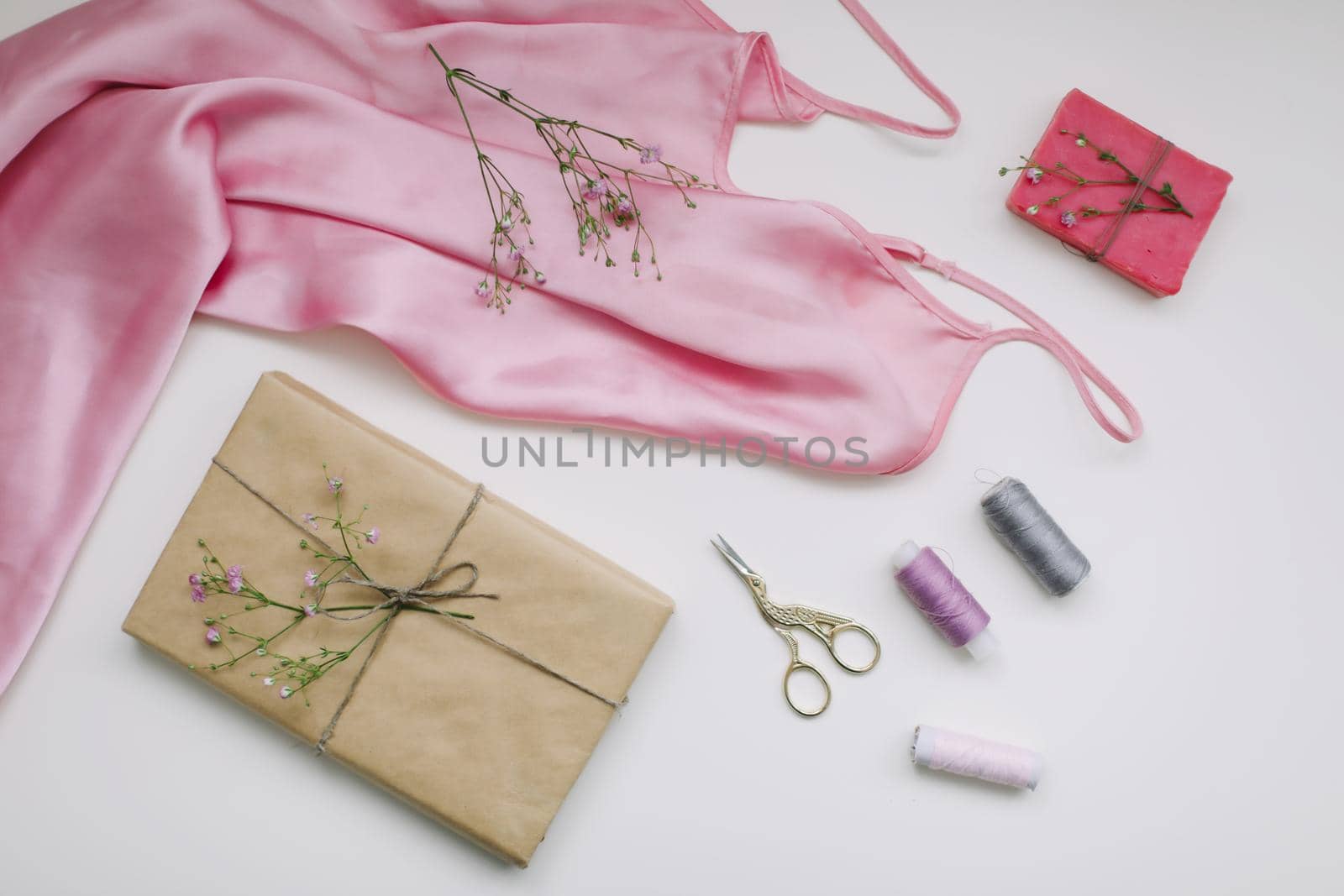 Composition with pink silk fabric, gift in craft paper and threads and scissors on white background. Flat lay, top view. Hobby, leisure concept