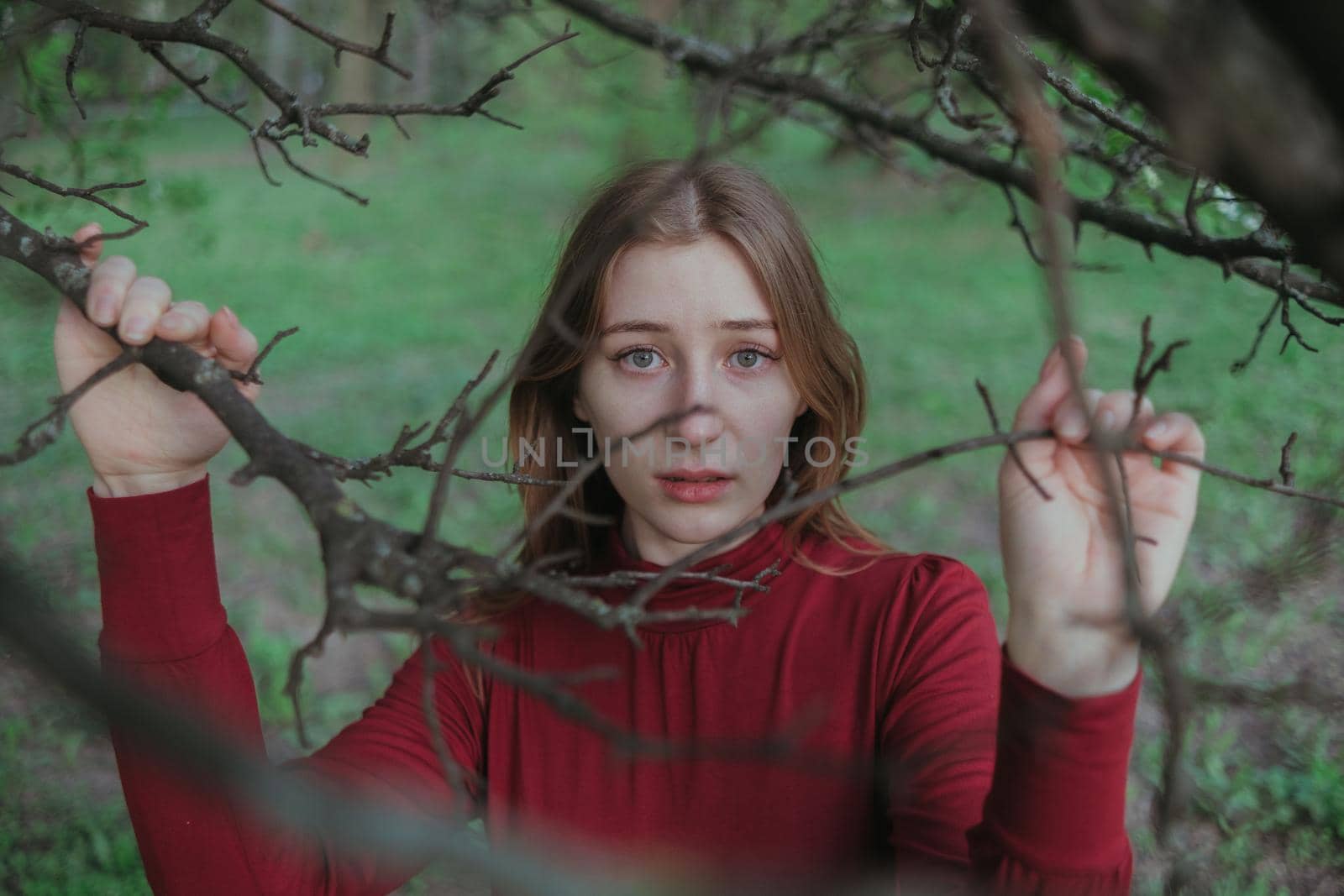 a blonde girl in red looks into the camera through the branches of a tree by Symonenko