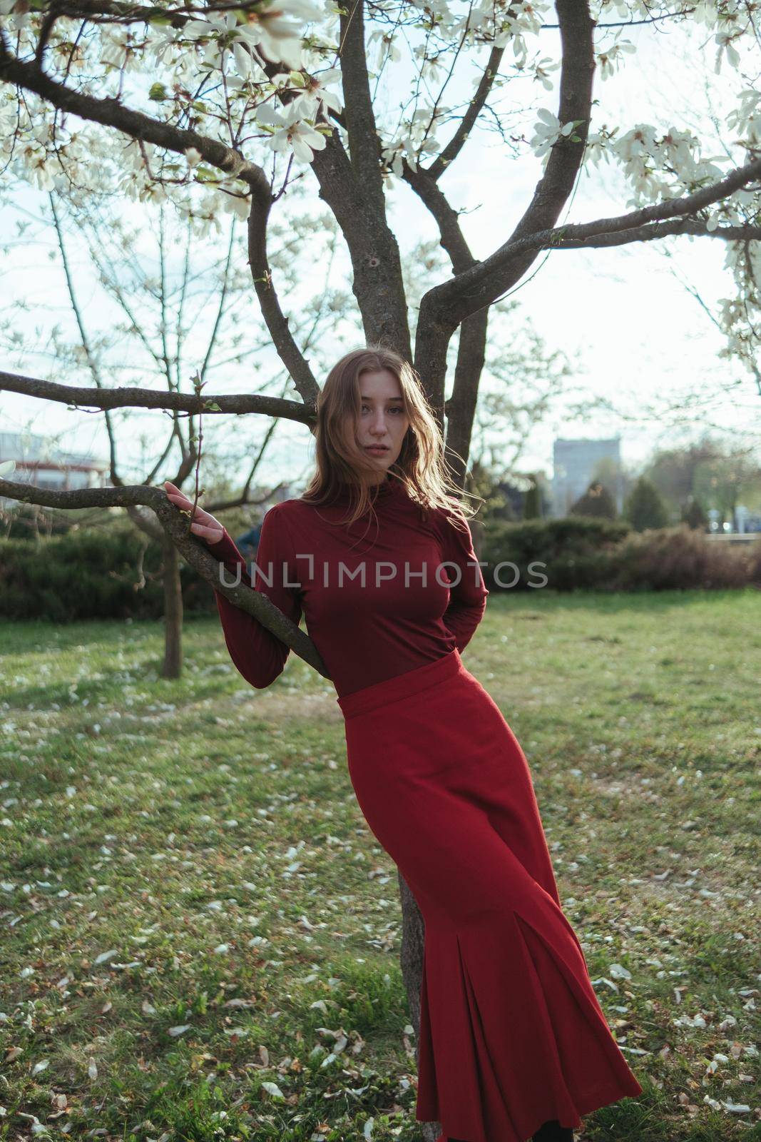 A blonde girl in red leans on a tree in the park. the concept of unity with nature