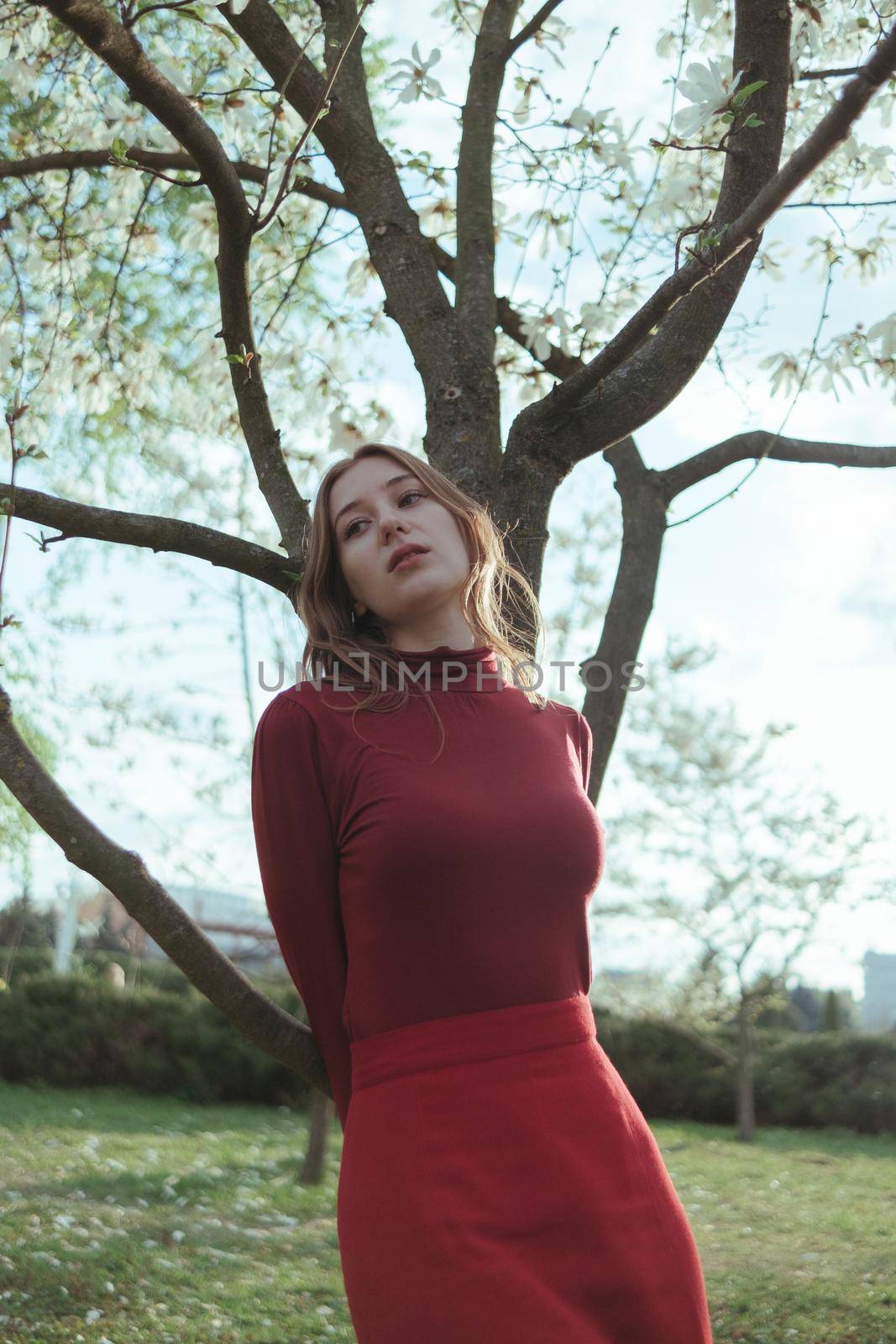 A blonde girl in red leans on a tree in the park. the concept of unity with nature