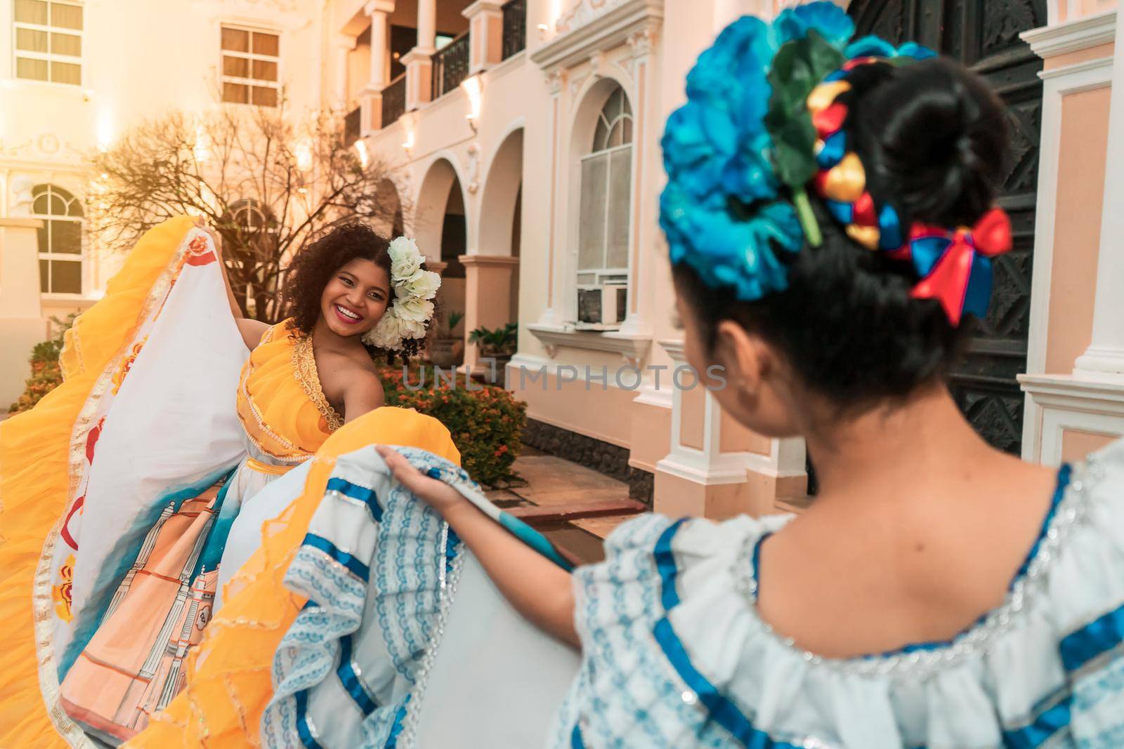 Two young Latina women, one mestizo and one Hispanic, dancing in traditional Nicaraguan costumes outside a colonial house at sunset.