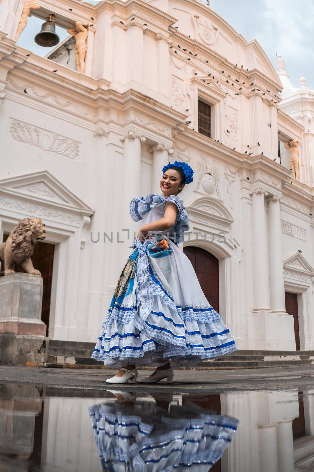 Vertical photo of a traditional dancer from Nicaragua in front of a puddle where you can see her reflection and the cathedral church of Leon de Nicaragua in the background..