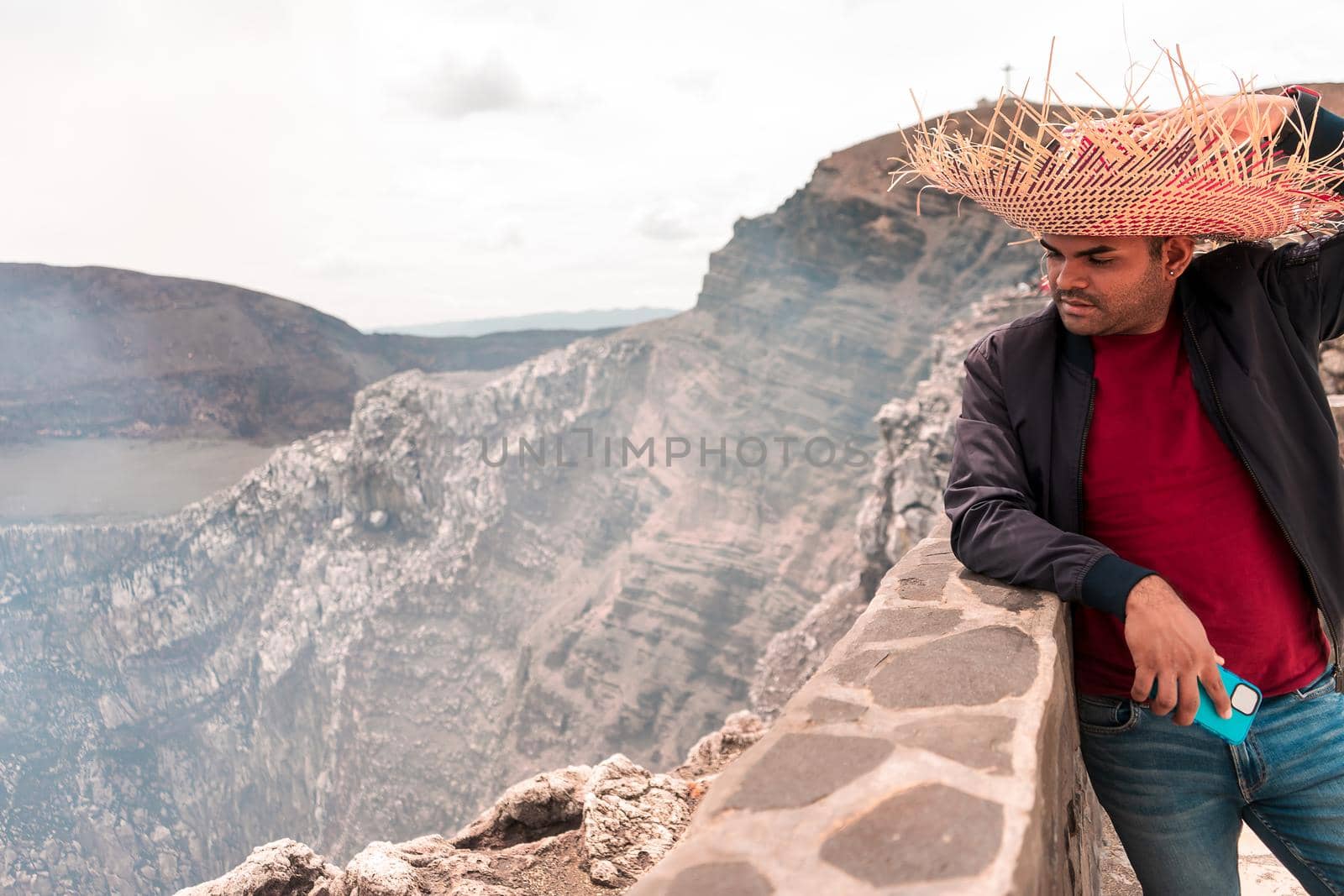 Mestizo latin man taking a selfie with his smartphone wearing a colorful hat looking towards the crater of the Masaya volcano in Nicaragua. Adventure and travel photo with copy space.