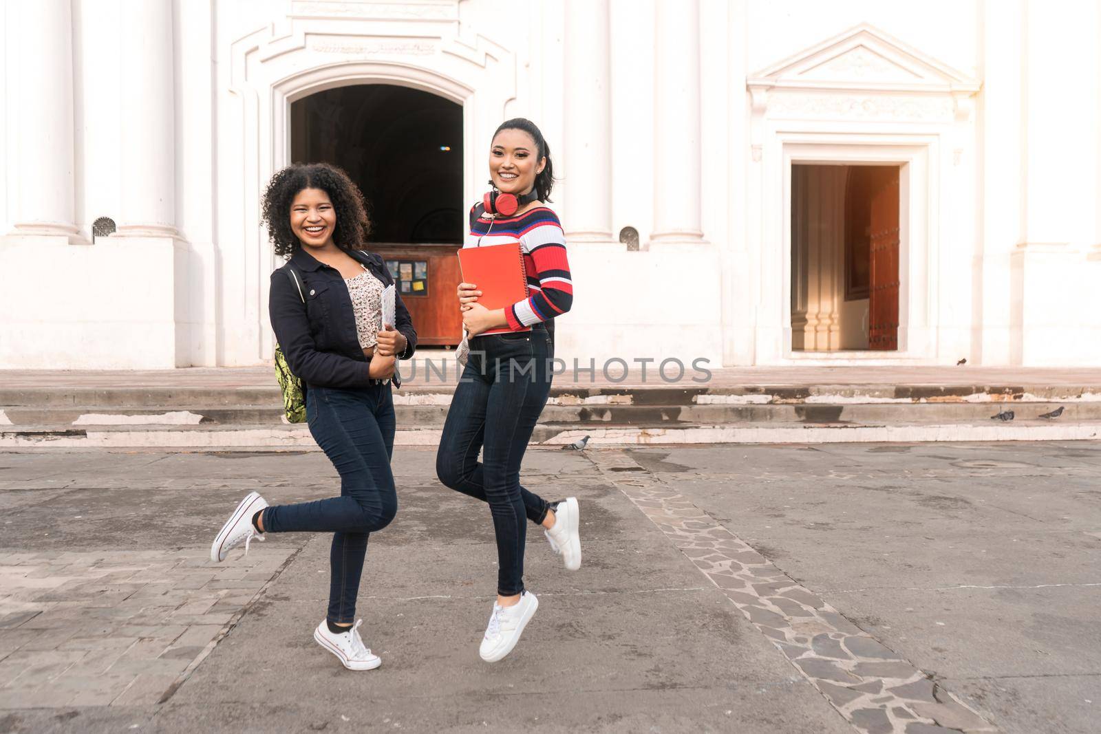 Two latin students jumping, one mestizo and one hispanic looking at camera and smiling holding notebooks in their hands and backpack on their backs. Behind them is the cathedral of Leon Nicaragua