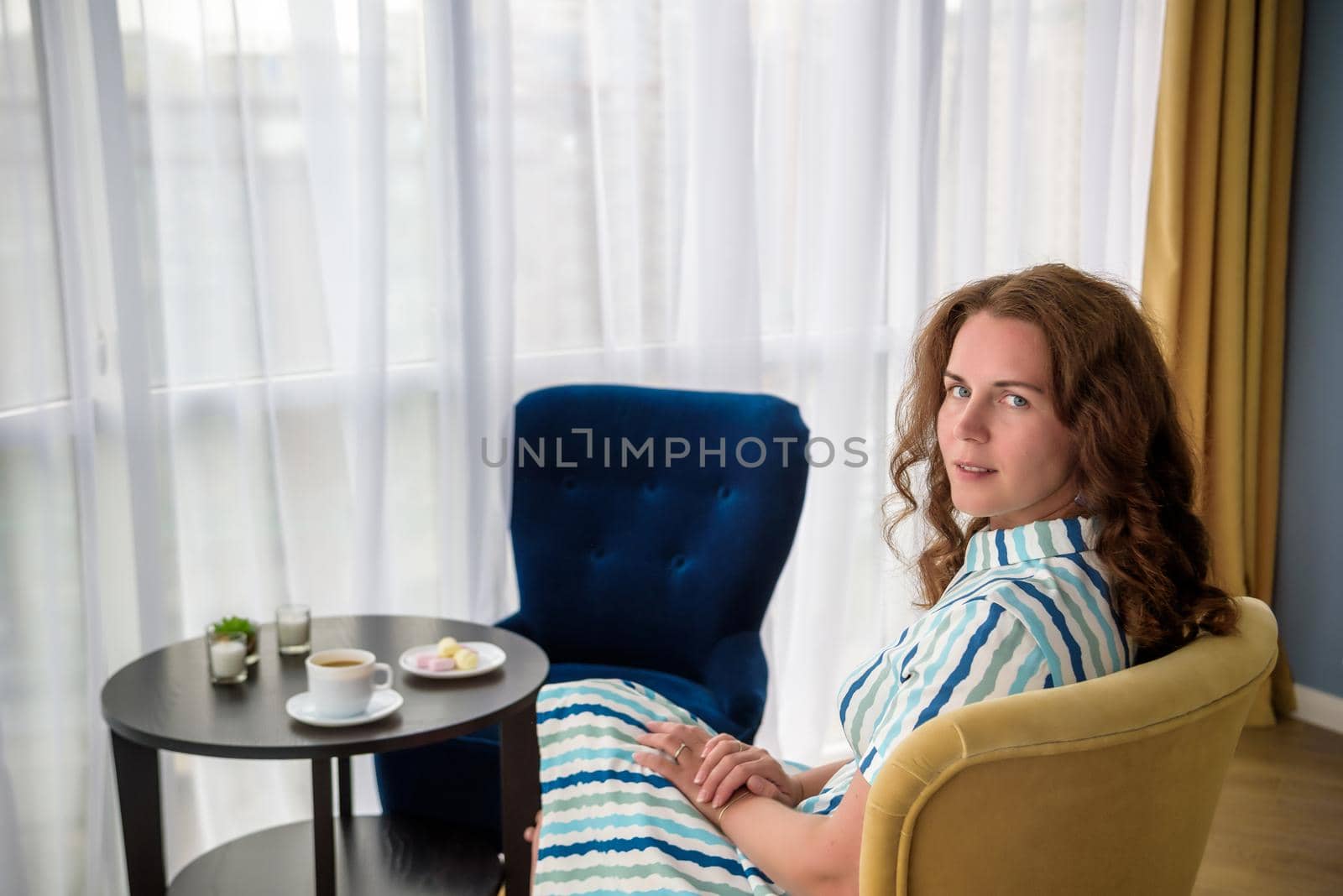 Young woman at home sitting on modern chair in front of window relaxing in her living room and drinking coffee or tea with sweets or desert. Relax before new business day.