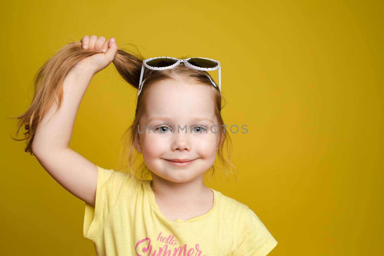 Front view of sunny little girl keeping hair in hand, and pointing with finger at copy space on yellow isolated background. Beautiful child with glasses looking at camera, smiling and posing.