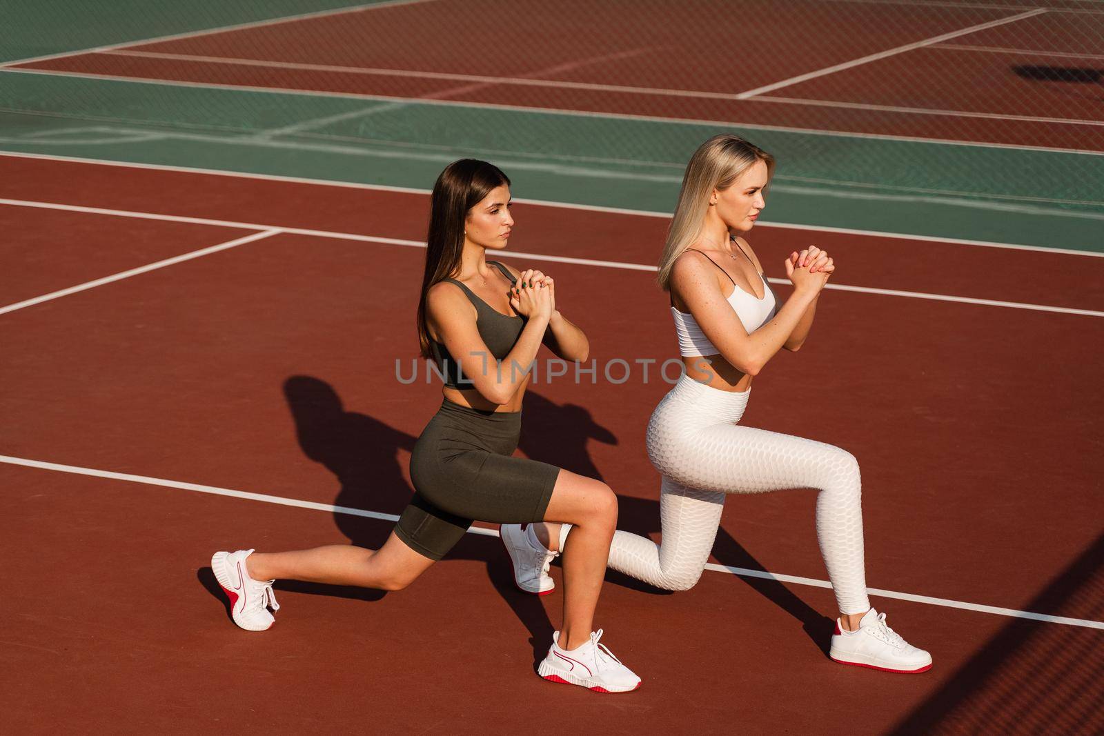 2 girls doing lunges training for stretching legs. Sport exercises on the tennis court