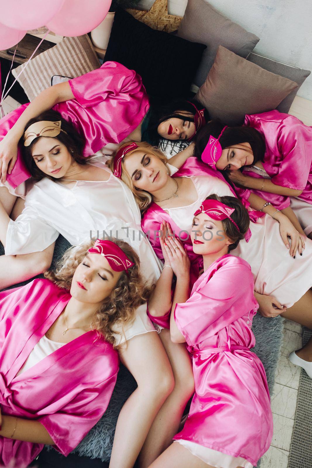 Overhead of pretty young ladies in pink bathrobes lying on bed with bride-to-be in white dress. Girlfriends showing tongues out and smiling. Bachelorette party.