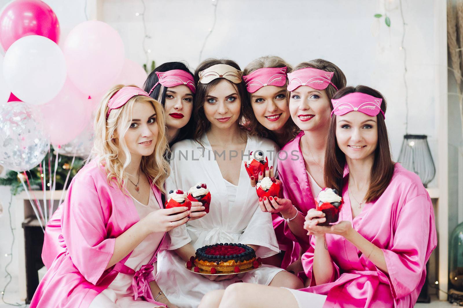 Portrait of beautiful young bridesmaids in pink robes and sleep masks holding cupcakes with berries. Attractive bride in white robe with berry cake.