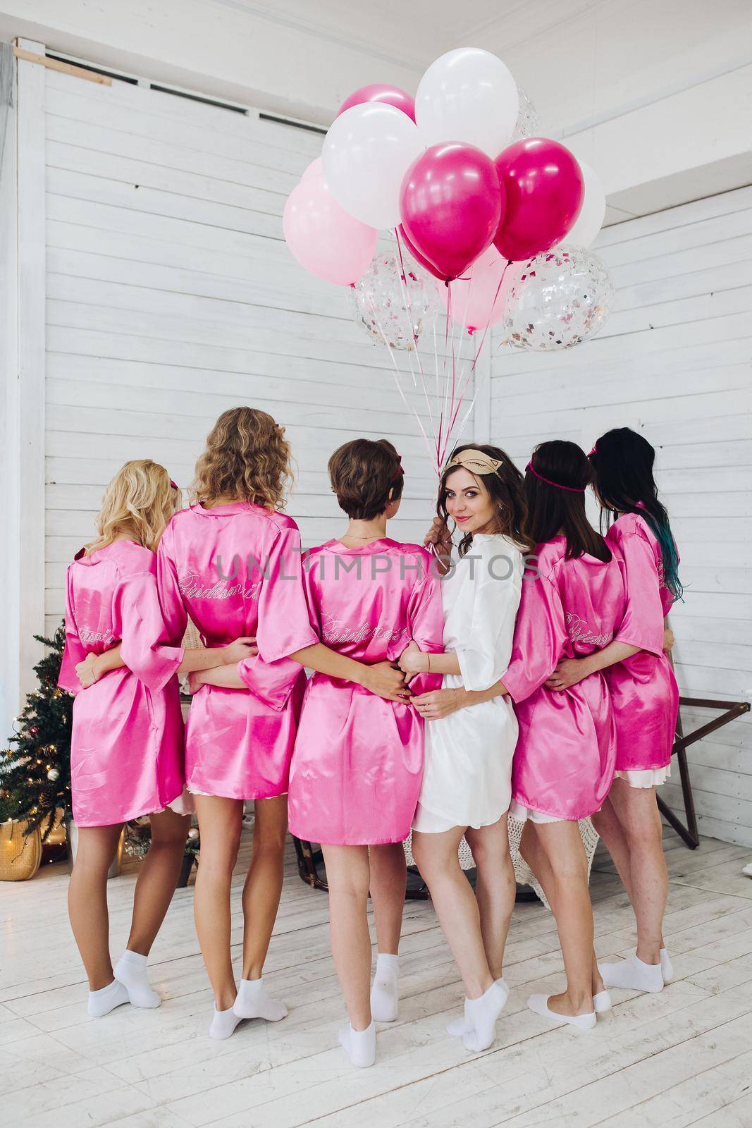 Studio shot of bride-to-be in white robe standing with her bridesmaids in pink robe back to camera with bunch of air balloons.