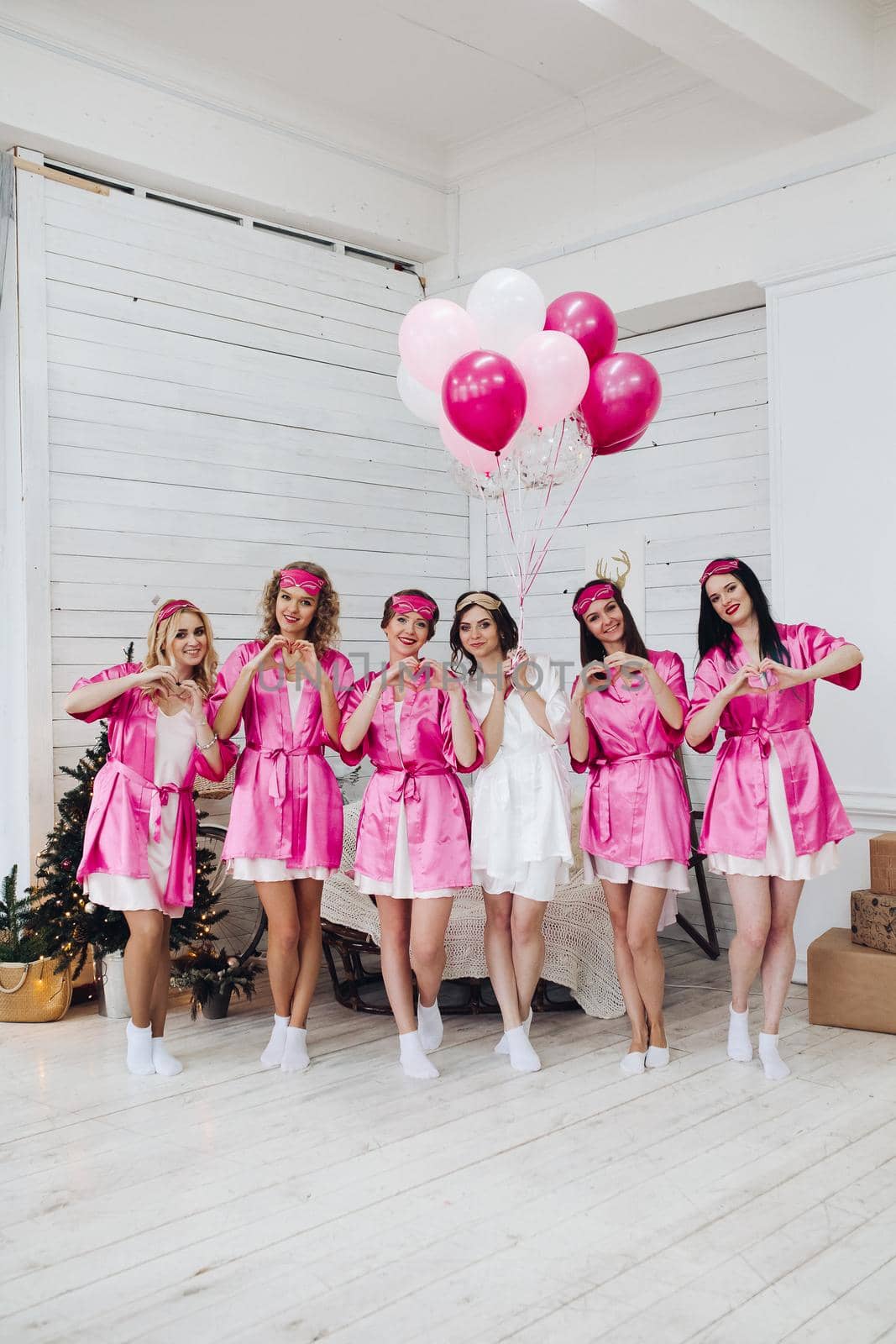 Full length portrait of happy lovely girlfriends in pink robes and sleeping masks jumping and having fun at hen party. Bride-to-be in white robe holding pink air balloons with smile standing on tiptoes.