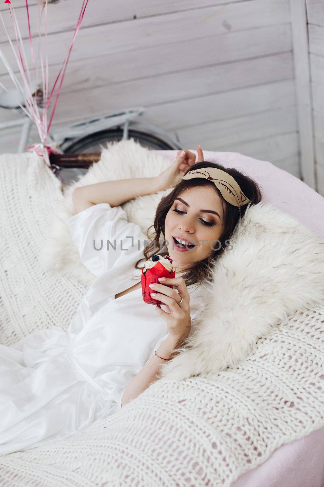 Portrait of attractive bride-to-be in white silk robe with sleeping mask on head holding delicious berry cake while relaxing in cozy white armchair.