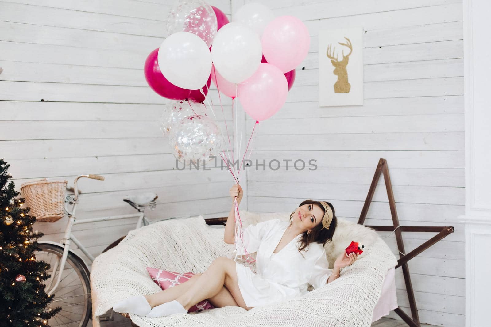 Portrait of pretty brunette bride-to-be in sleeping mask and white robe with cupcake and bunch of pink cute air balloons relaxing in cozy round chair.