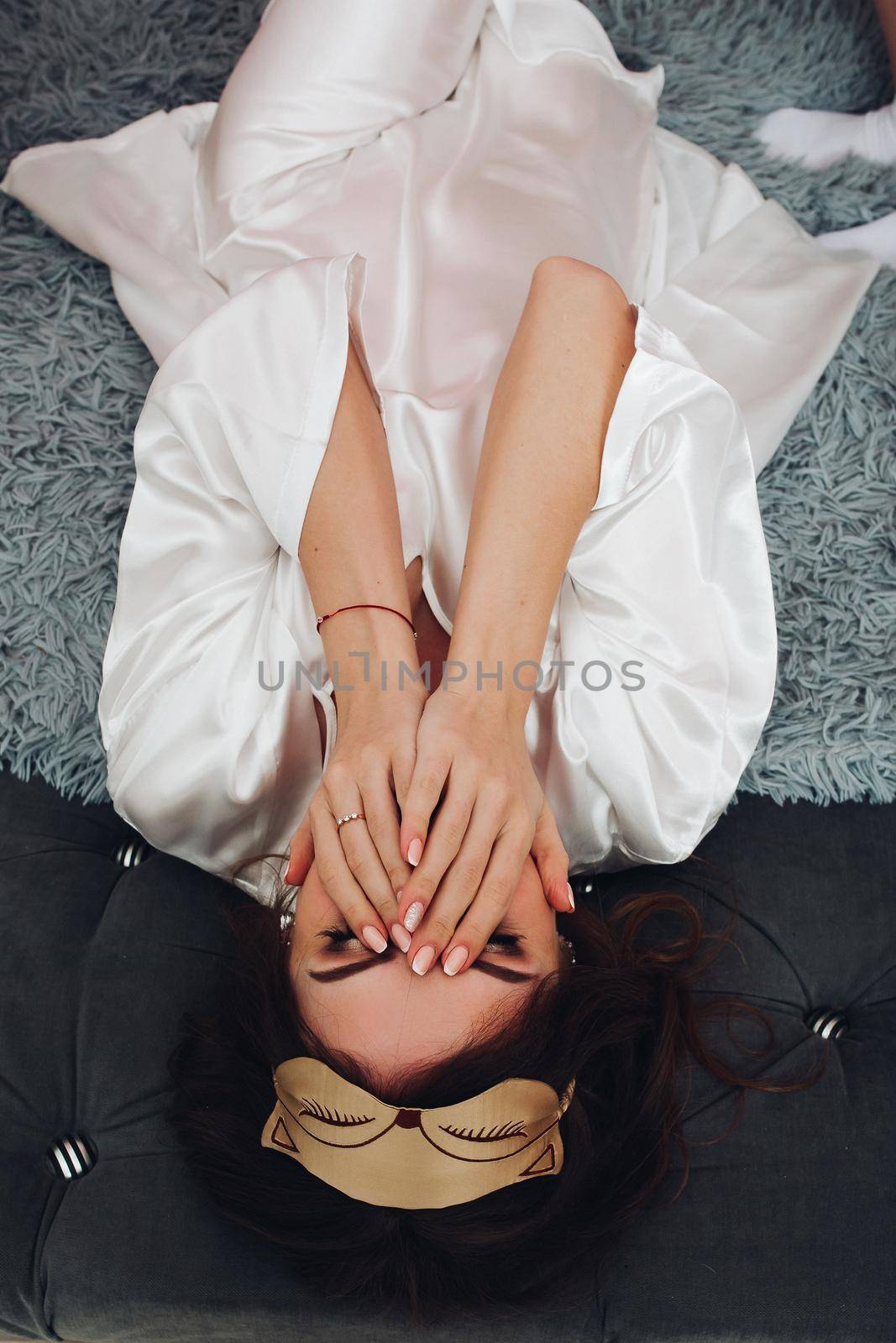 Top view of brunette young girl in white silk robe and cat sleeping mask covering face with hands lying on bed.