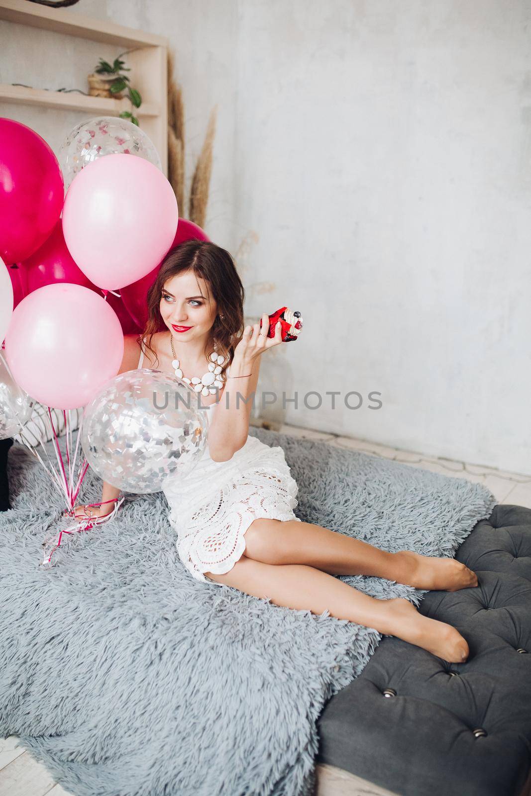 Portrait of pretty young bride-to-be in white dress sitting on sofa with muffin and pink air balloons. Hen party.