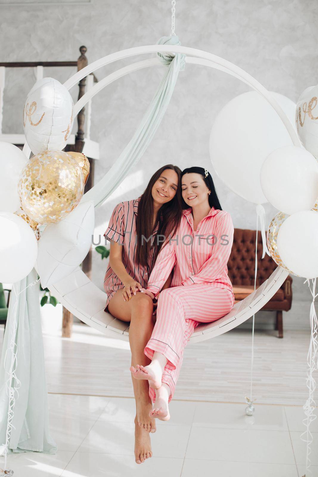 Full length of two beautiful girls in pink striped pyjamas sitting barefoot snuggling on white swings decorated with air balloons. Bride-to-be concept. Hen party with the best friend.