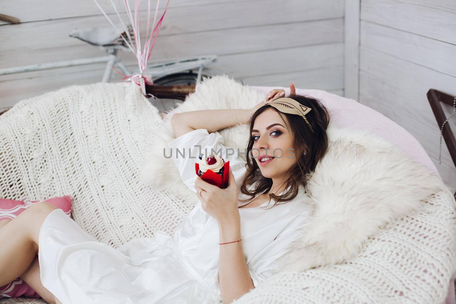 Portrait of attractive bride-to-be in white silk robe with sleeping mask on head holding delicious berry cake while relaxing in cozy white armchair.