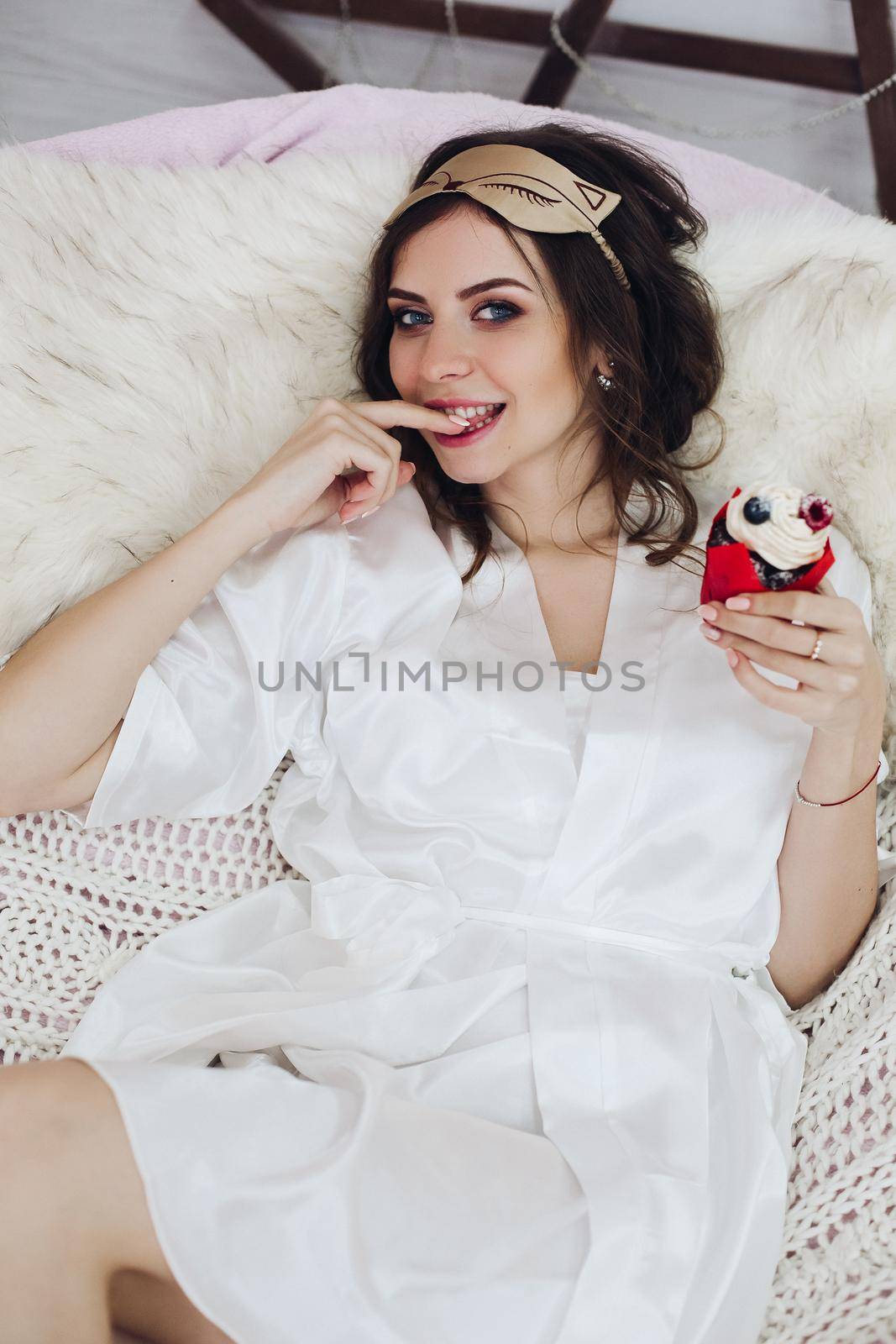 Portrait of pretty elegant bride-to-be in sleep mask and white silk robe relaxing in armchair holding berry muffin and flirting with camera.