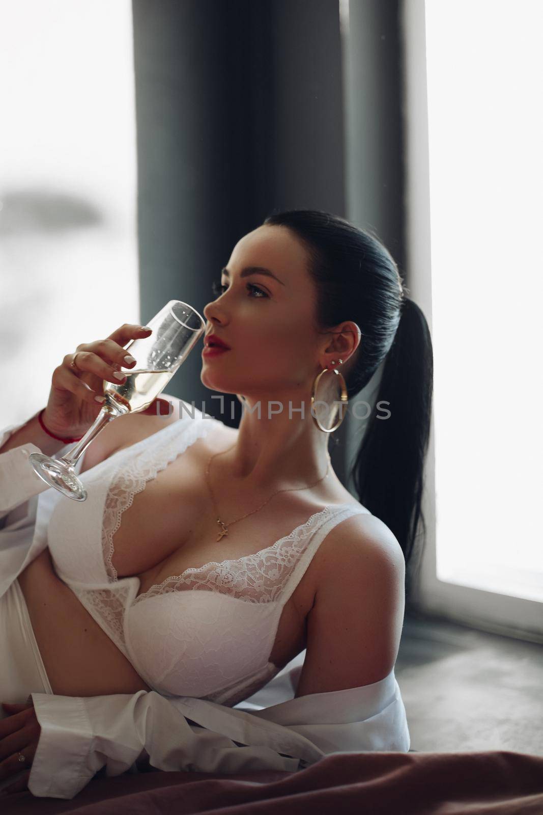 Fashion brunette girl in lingerie lying on bed with bokal of champagne enjoying luxury lifestyle by StudioLucky