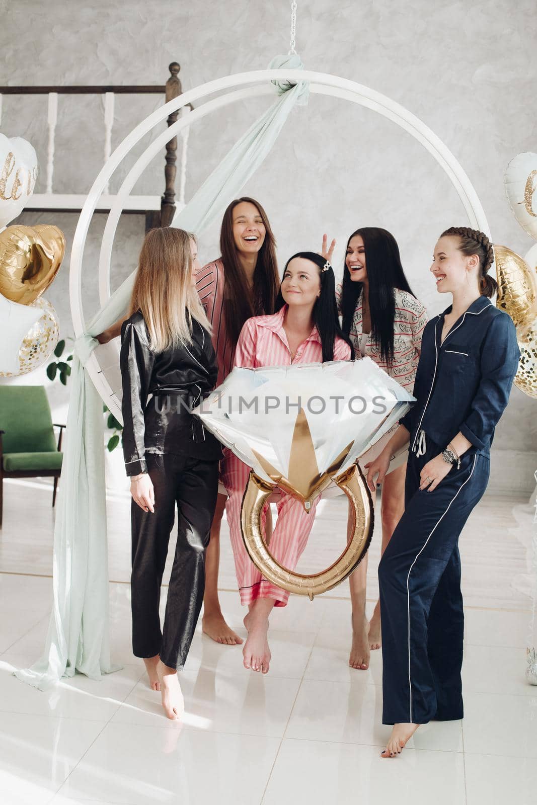 Five friends celebrating the party dedicated to a bachelorette in the middle. Hen party celebration. Girls laughing and smiling with black-haired girl in the middle holding a big balloon ring.