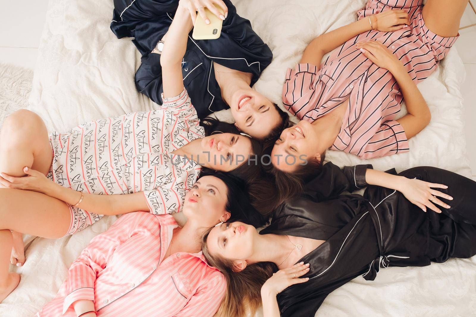 Top view stock photo of five beautiful girlfriends in lovely home apparel making selfie laying on cozy bed. Bride-to-be taking selfie with her mobile phone with her best friends or bridesmaids while resting on bed. They are making duck faces posing to camera.