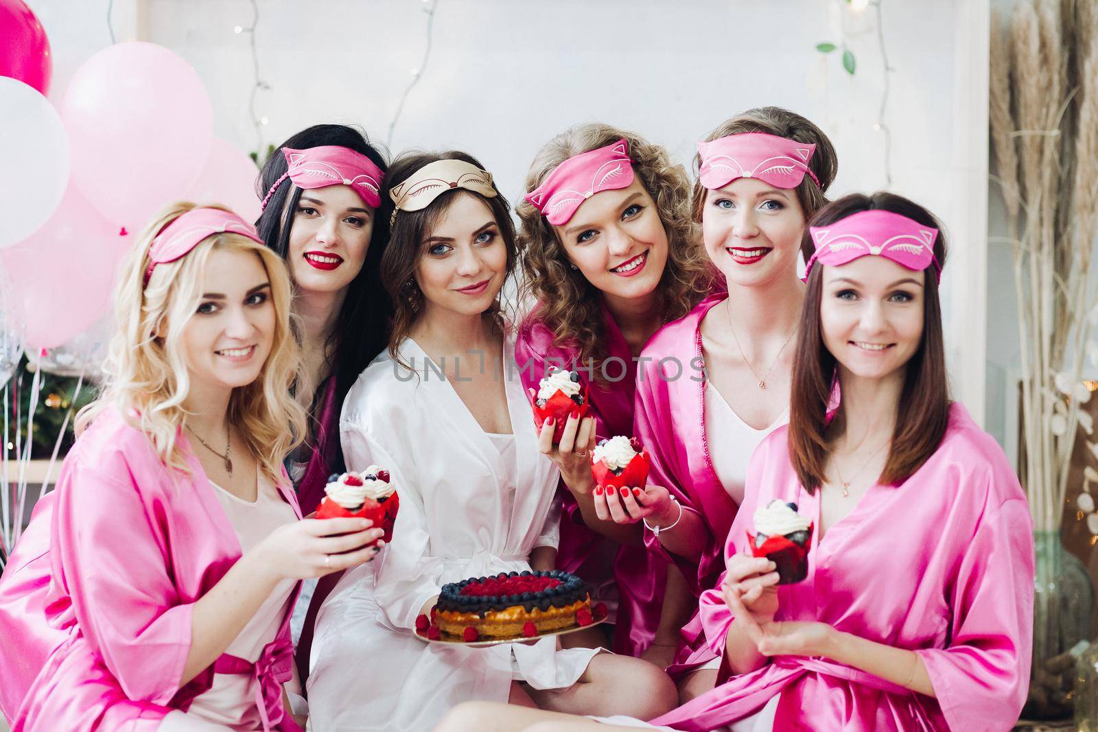 Portrait of beautiful young bridesmaids in pink robes and sleep masks holding cupcakes with berries. Attractive bride in white robe with berry cake.