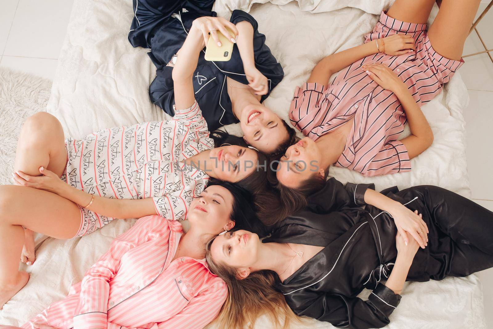 Top view stock photo of five beautiful girlfriends in lovely home apparel making selfie laying on cozy bed. Bride-to-be taking selfie with her mobile phone with her best friends or bridesmaids while resting on bed. They are making duck faces posing to camera.