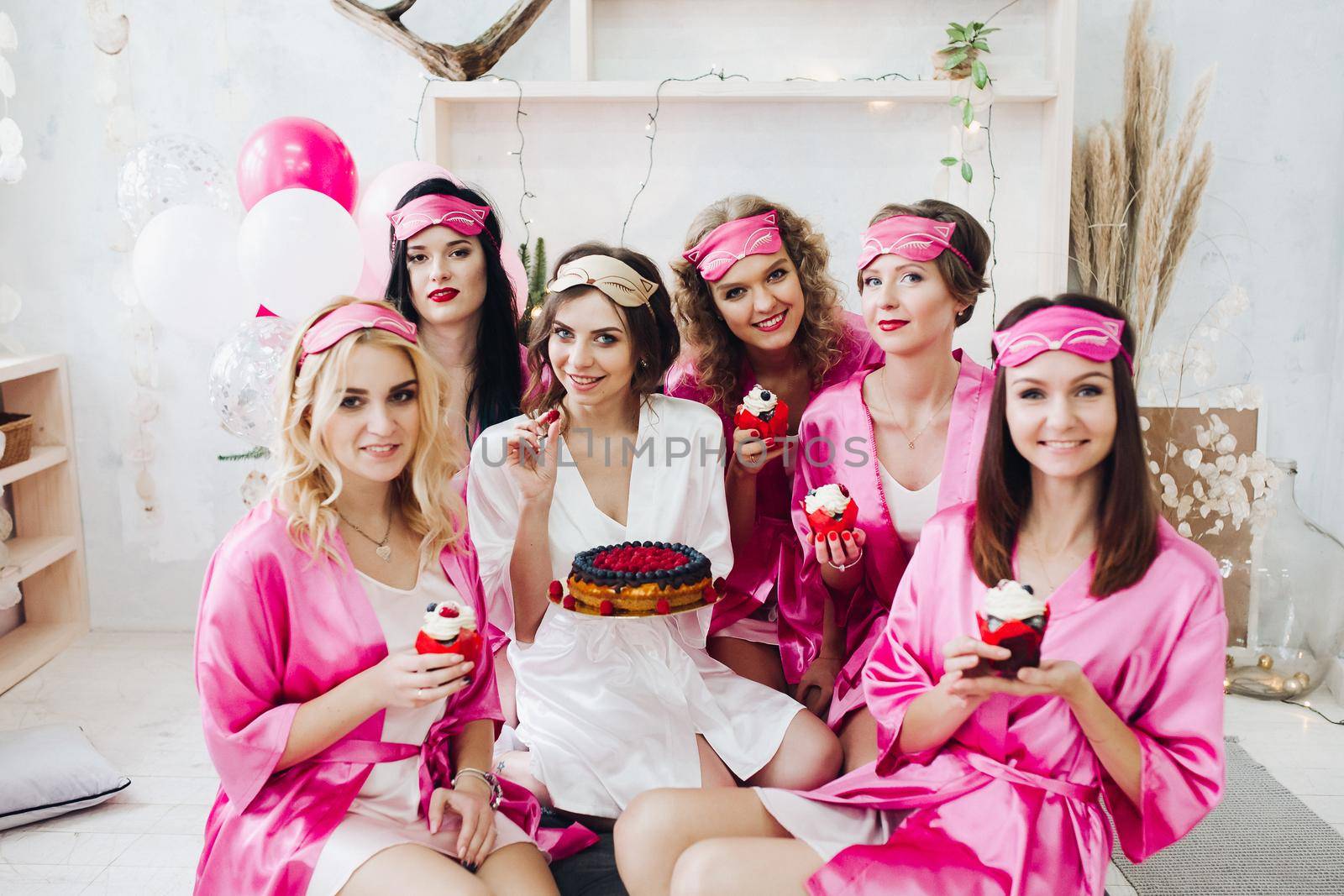 Portrait of beautiful young bridesmaids in pink robes and sleep masks holding cupcakes with berries. Attractive bride in white robe with berry cake.