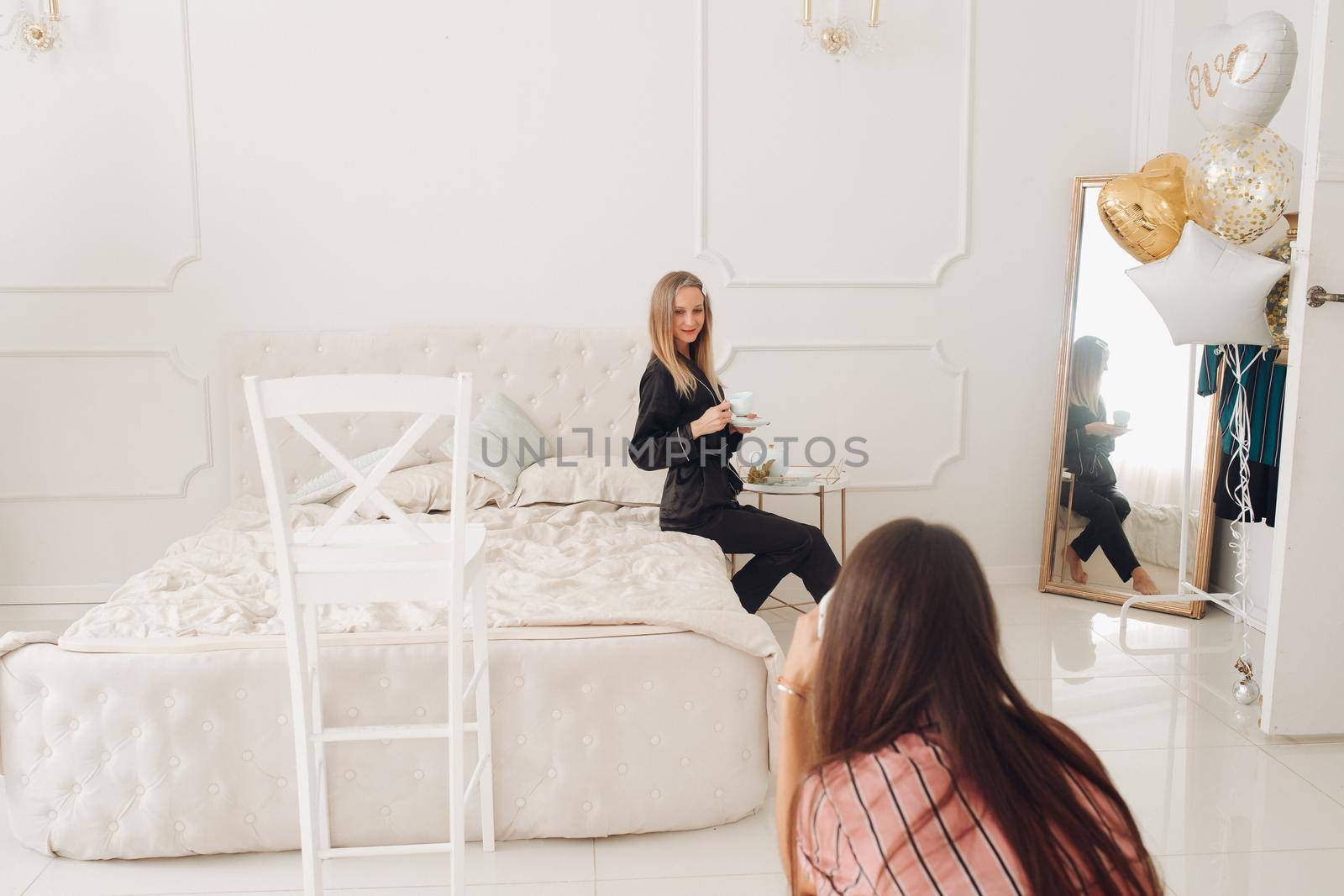 A friend taking photo of gorgeous blonde woman in elegant black pyjamas sitting on white bed with cup of morning coffee in beautiful white room. There are air balloons next to the mirror.