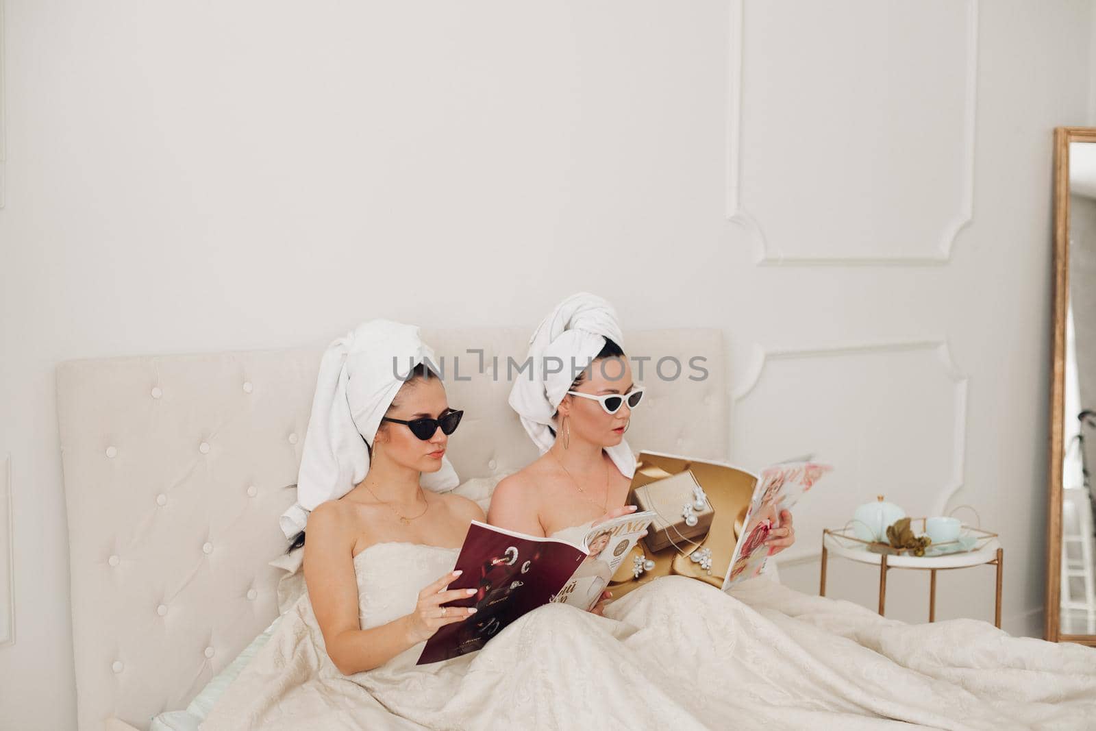 Young females in sunglasses and towels sitting on bed while reading magazines. Care, leisure and relaxation time
