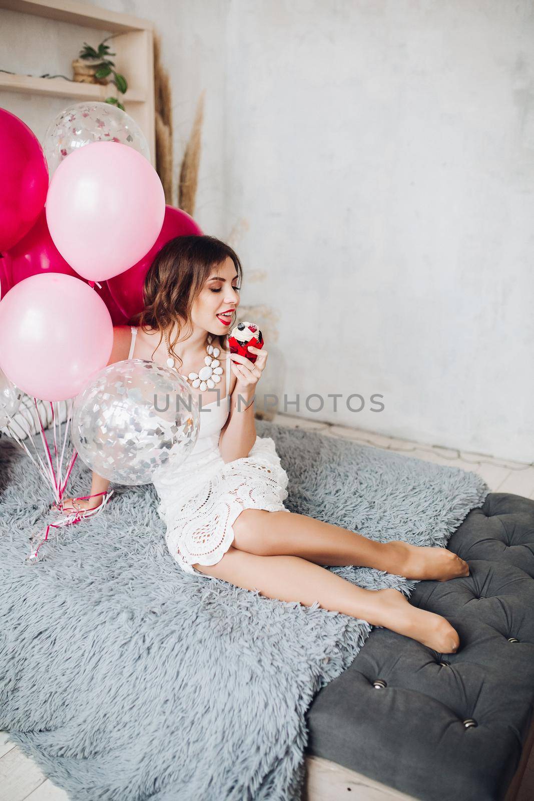 Portrait of pretty young bride-to-be in white dress sitting on sofa with muffin and pink air balloons. Hen party.