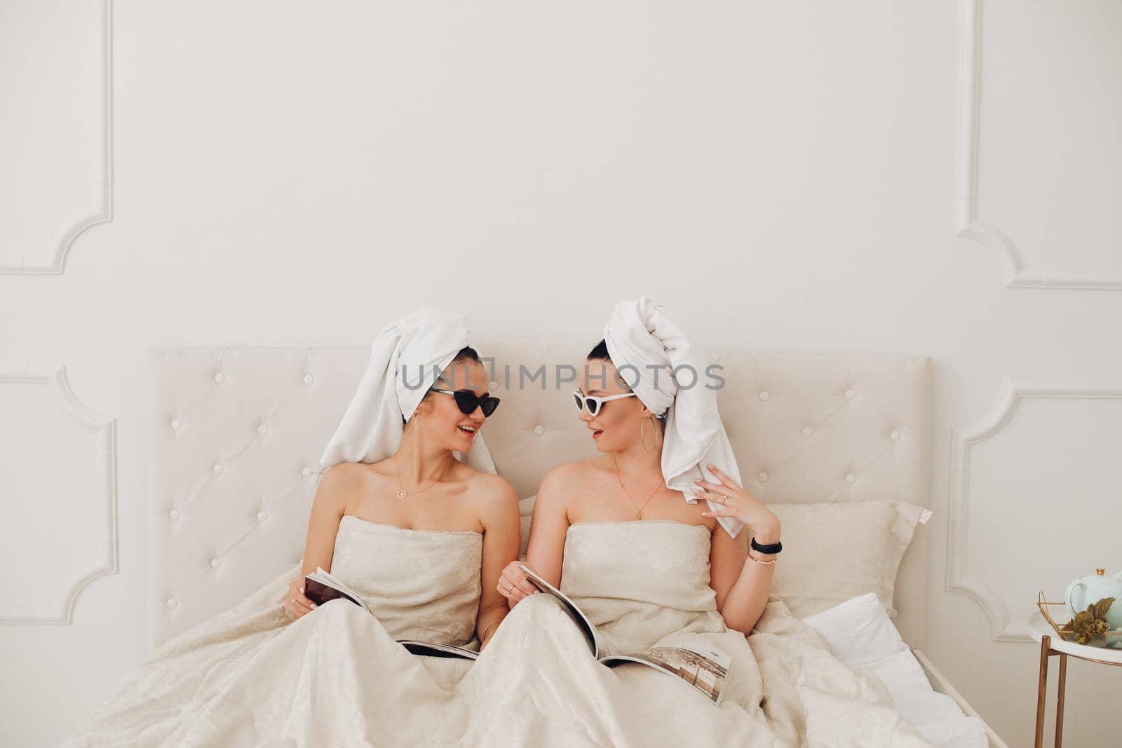 Young females in sunglasses and towels sitting on bed while reading magazines. Care, leisure and relaxation time