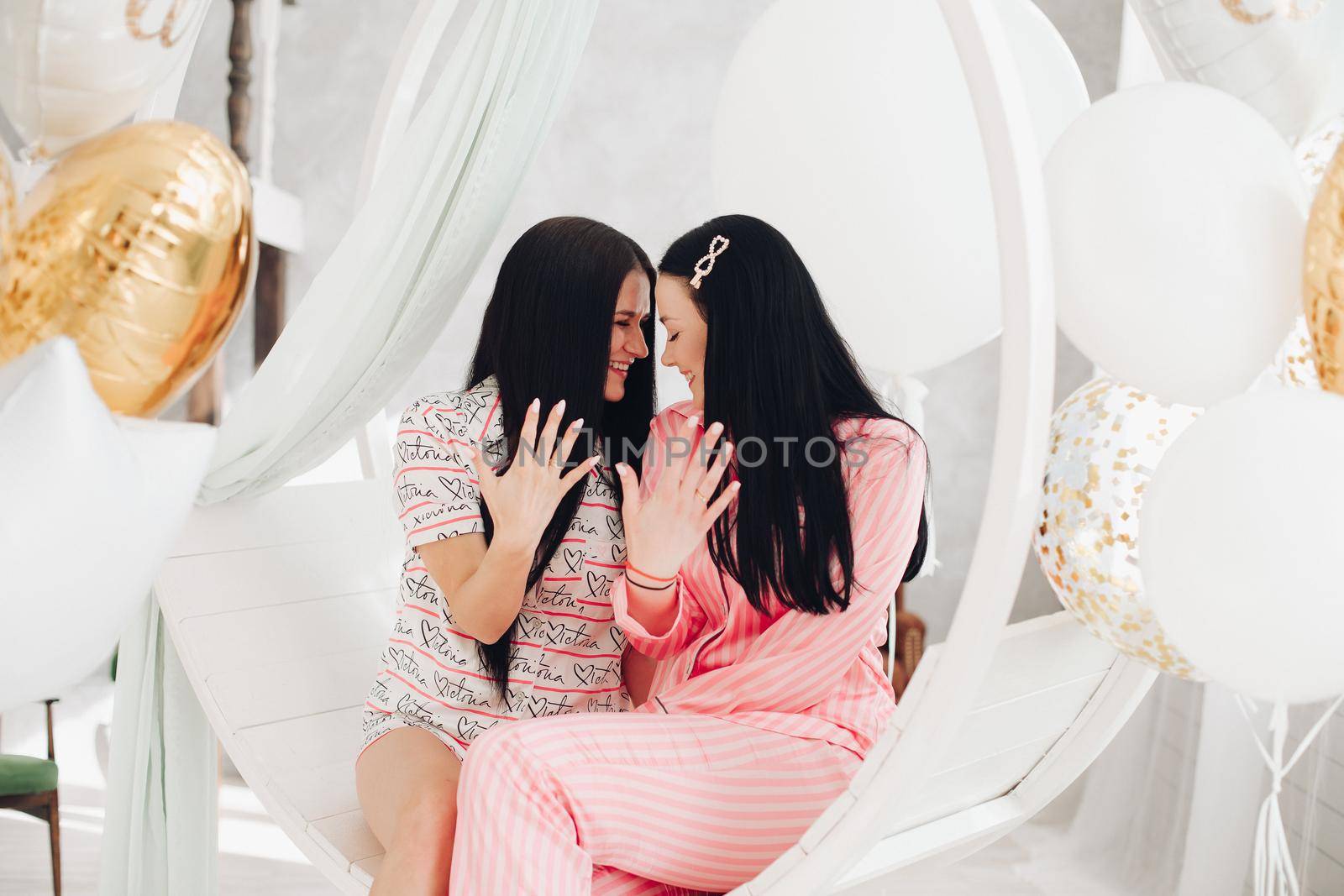 Two happy female friend in pajamas sitting on stylish armchair at luxury interior. Smiling girlfriends enjoying friendship surrounded by festive party event having positive emotion