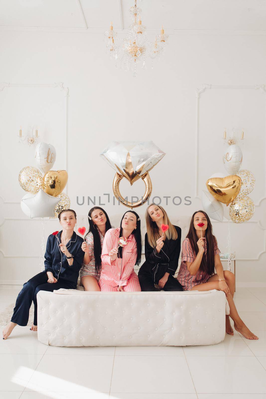 Stock photo of five girls in pajamas eating sweet lollipops sitting close on big white bed in beautiful white room with inflated air balloons in shape of wedding ring, hearts and stars.