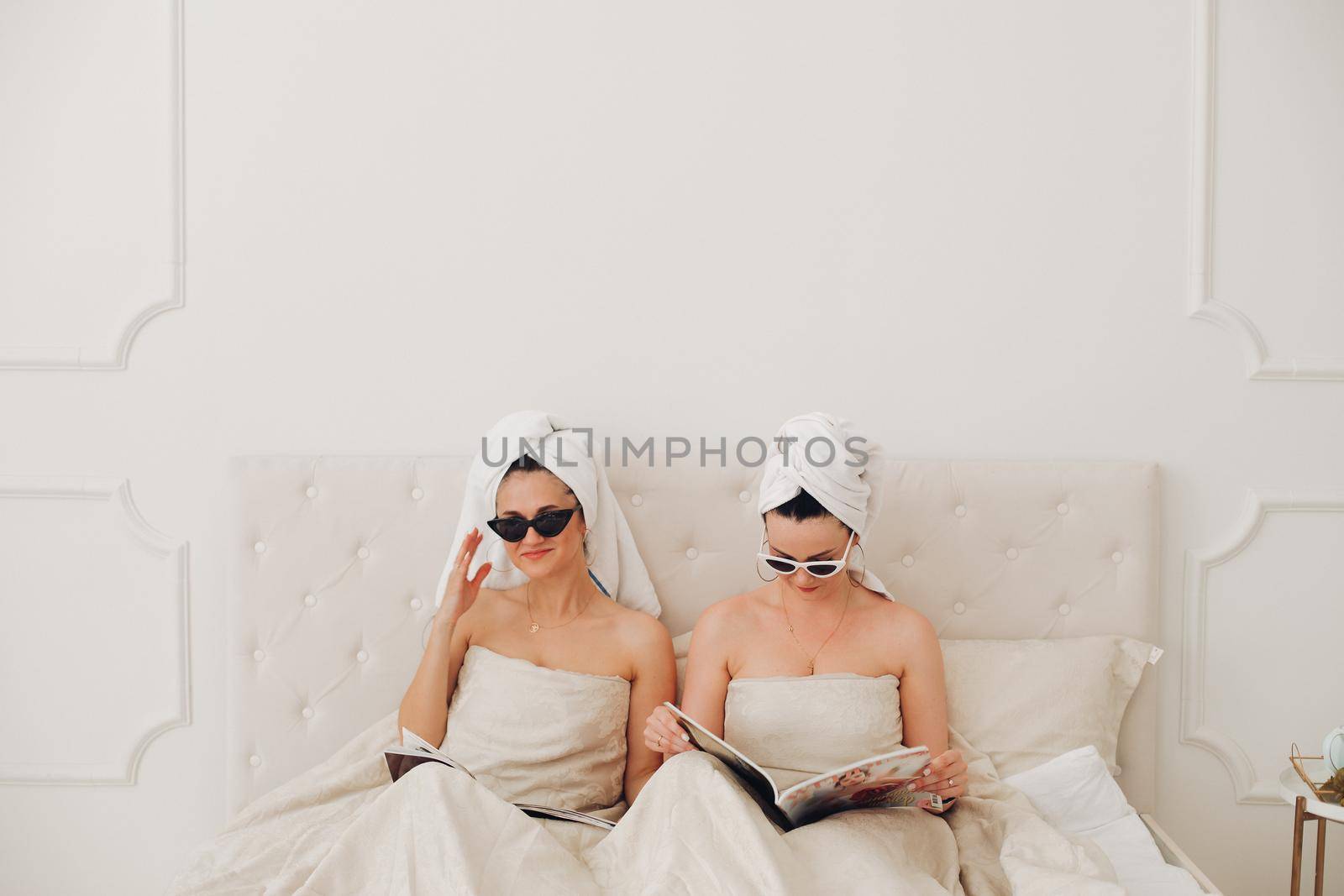 Stock photo of incognito women in bath towels on head and sunglasses holding fashion magazines sitting in bed. Fashion victims in the morning. Fashion divas preparing for the day reading fashion journals in bed.