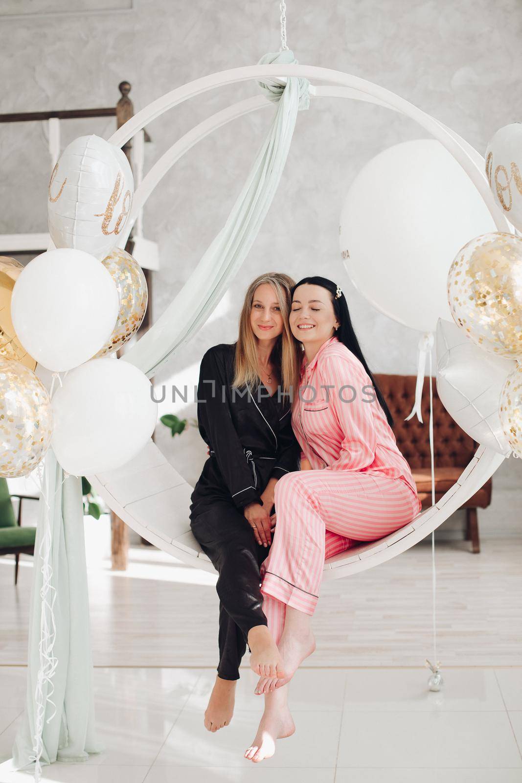 Two happy female friend in pajamas sitting on stylish armchair at luxury interior by StudioLucky