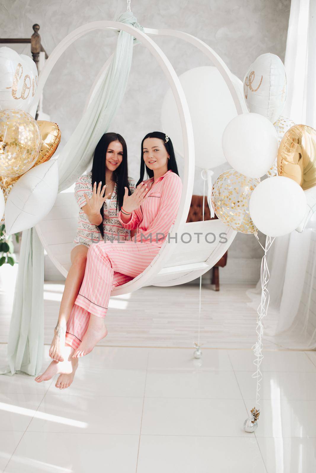 Two happy female friend in pajamas sitting on stylish armchair at luxury interior. Smiling girlfriends enjoying friendship surrounded by festive party event having positive emotion