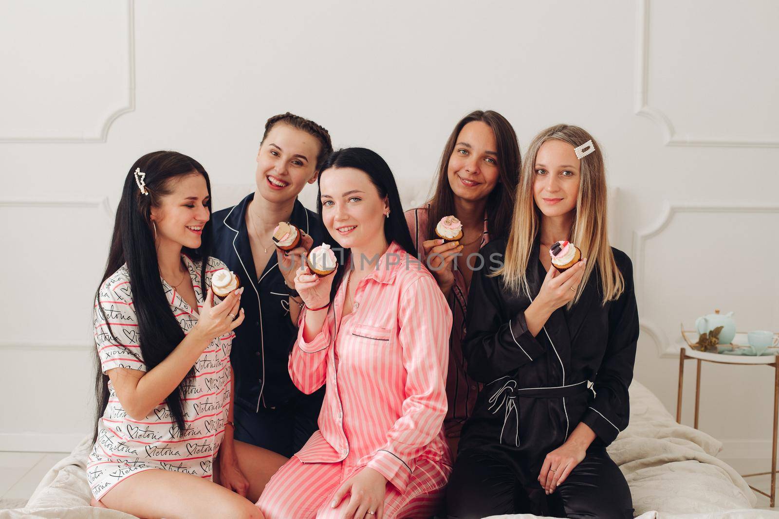 Happy pretty women in sleepwear sitting on the edge of the bed and holding cupcakes. Friendship and happiness concept