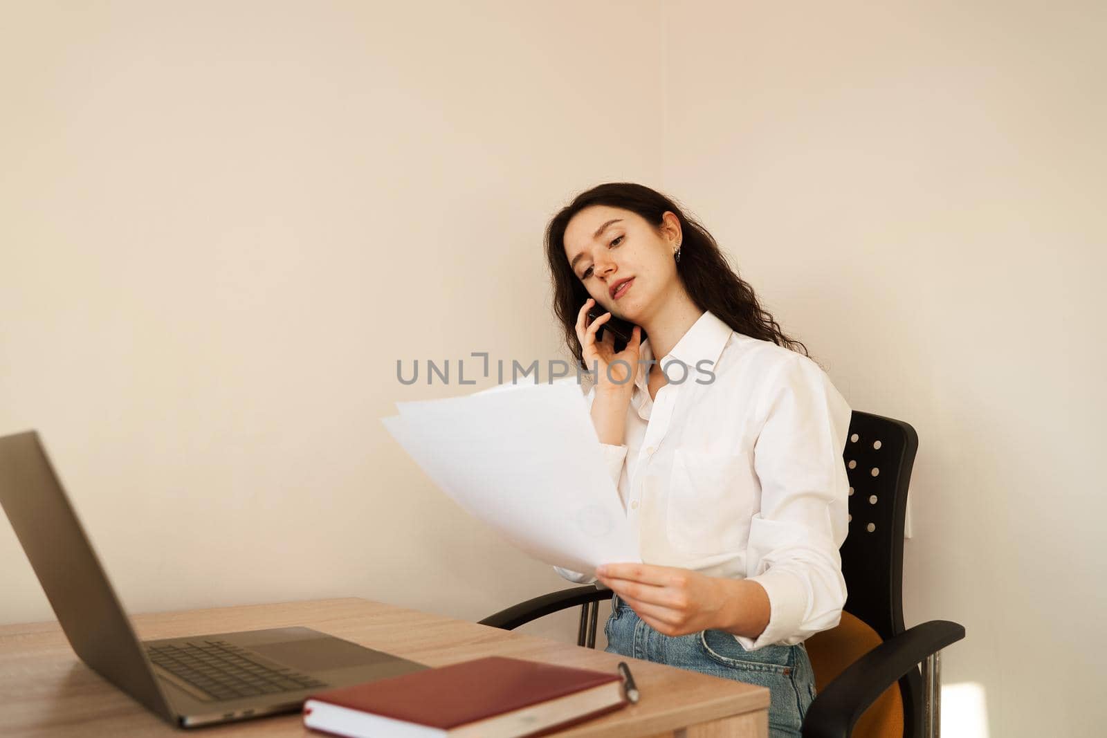 Cheerful woman manager talking by phone, doing paperwork and working on laptop. Girl boss work in the office