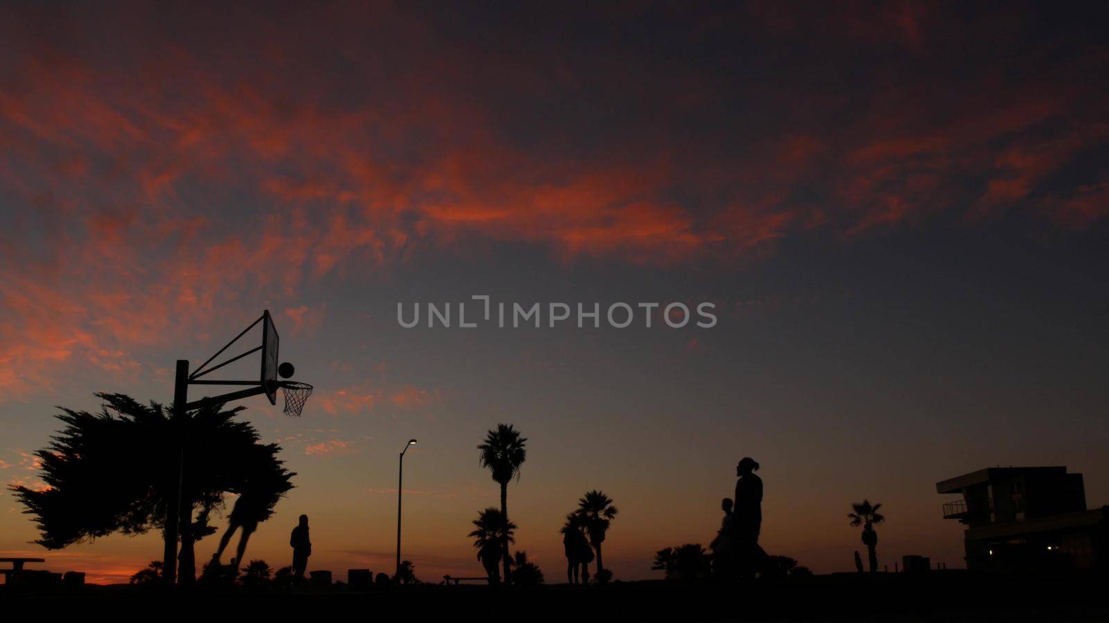 People playing basket ball game, silhouettes of players on basketball court outdoor, sunset ocean beach, California coast, Mission beach, USA. Black hoop, net and backboard on streetball sport field.