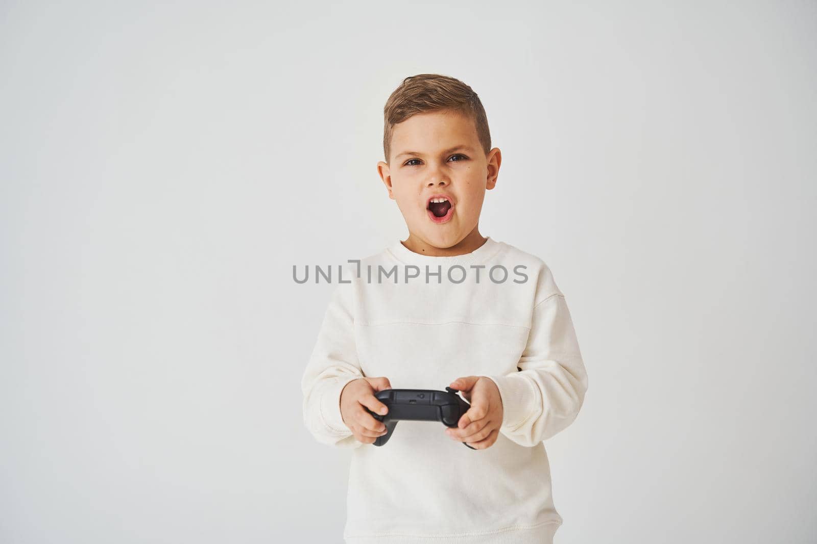 Surprised kid with gamepad playing console games on white background. Gambling addiction of child. by Rabizo