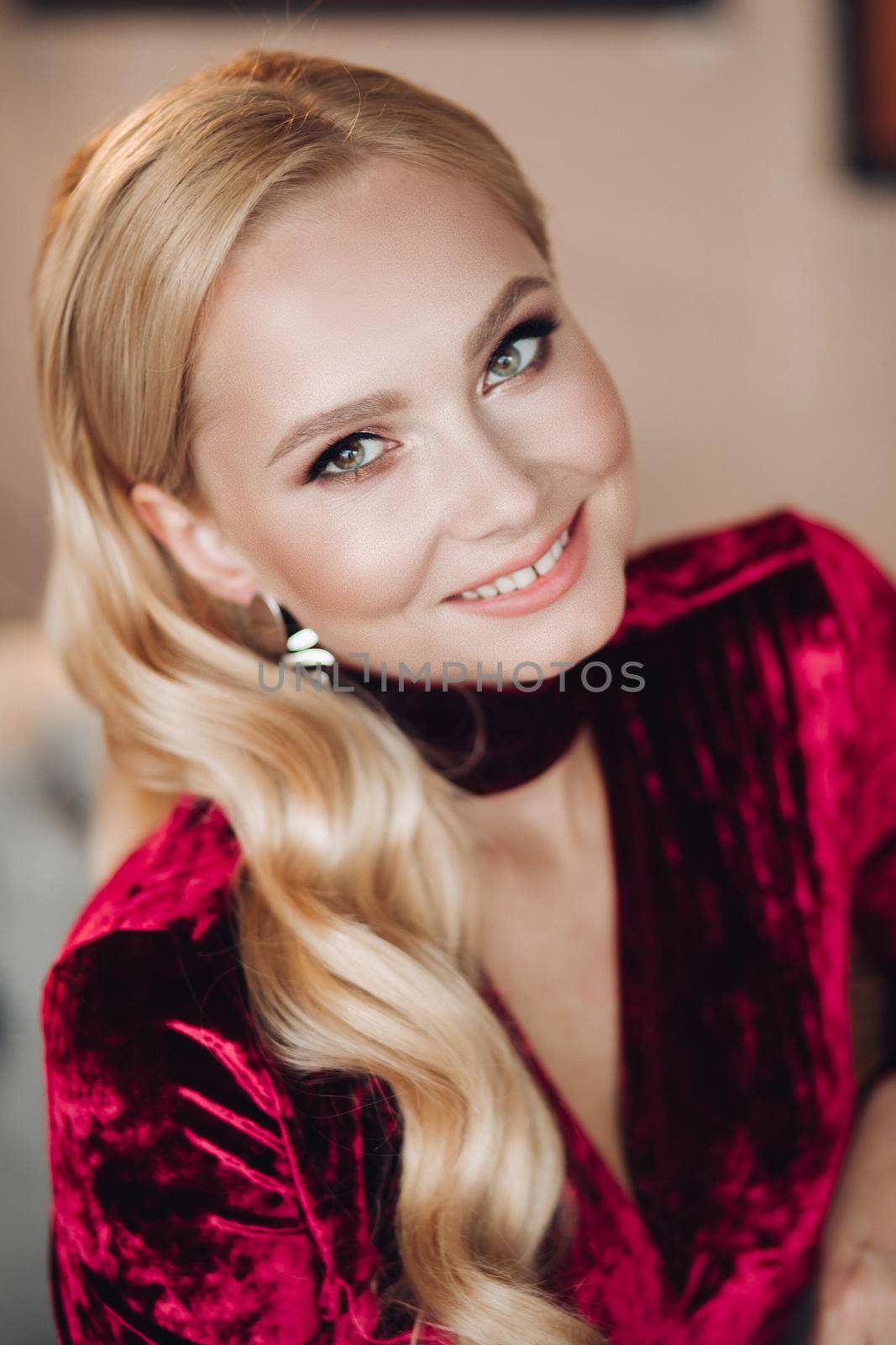 Close up from above of beautiful young woman in dark red dress looking at camera. Happy blonde girl with long hair with make up smiling. Portrait of gorgeous model sitting and posing in elegant look.