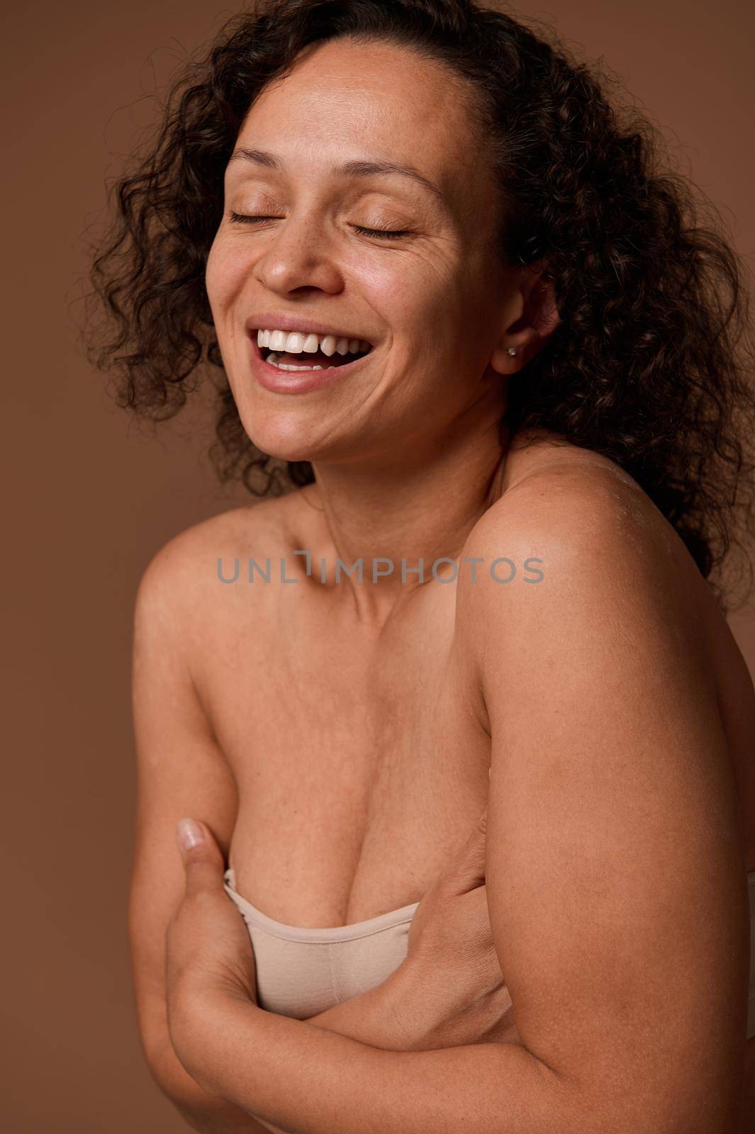 Delighted confident dark-haired curly woman with closed eyes and skin flaws in beige underwear smiles toothy smile posing against beige background with copy space for ads