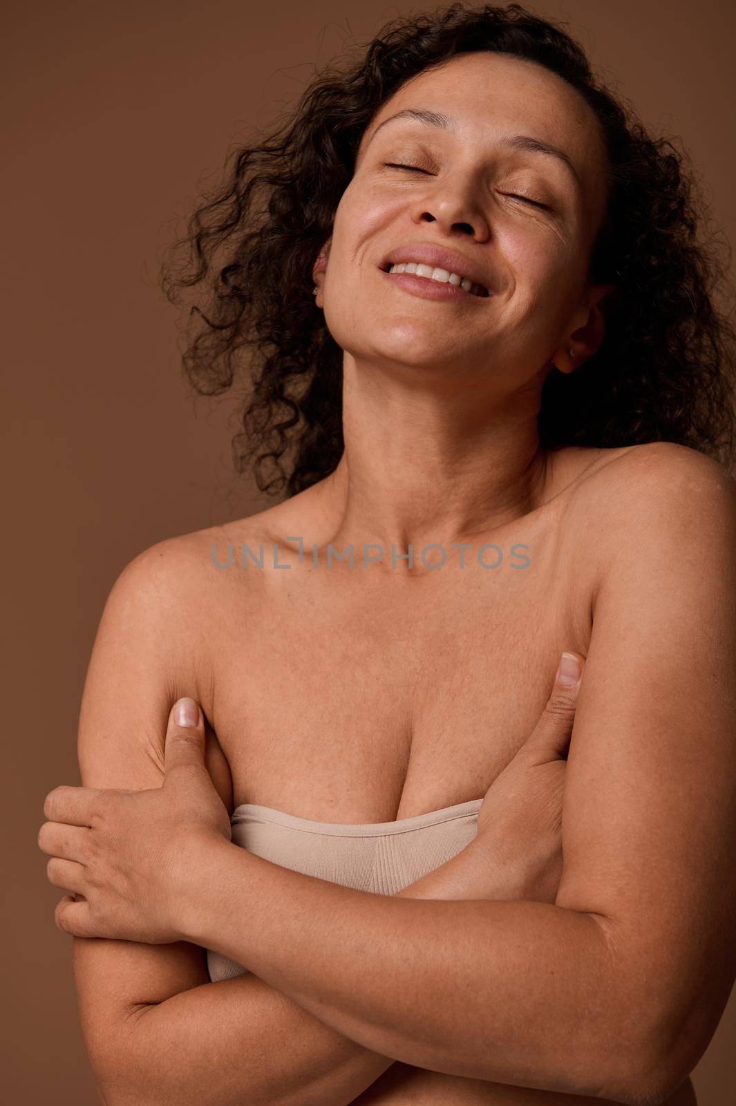 Delighted dark-haired curly woman with closed eyes and skin flaws and imperfections in beige underwear smiles toothy smile posing against beige background with copy space for ads