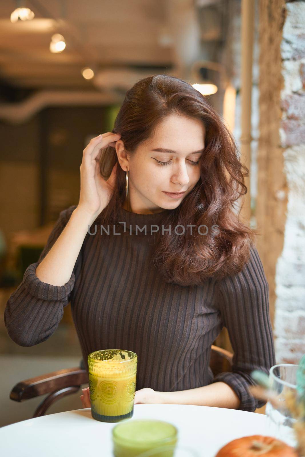 Beautiful serious stylish fashionable smart girl is sitting in cafe and drinking healthy yellow smoothie or latte vegan. Charming thoughtful woman with long dark brown hair. by NataBene