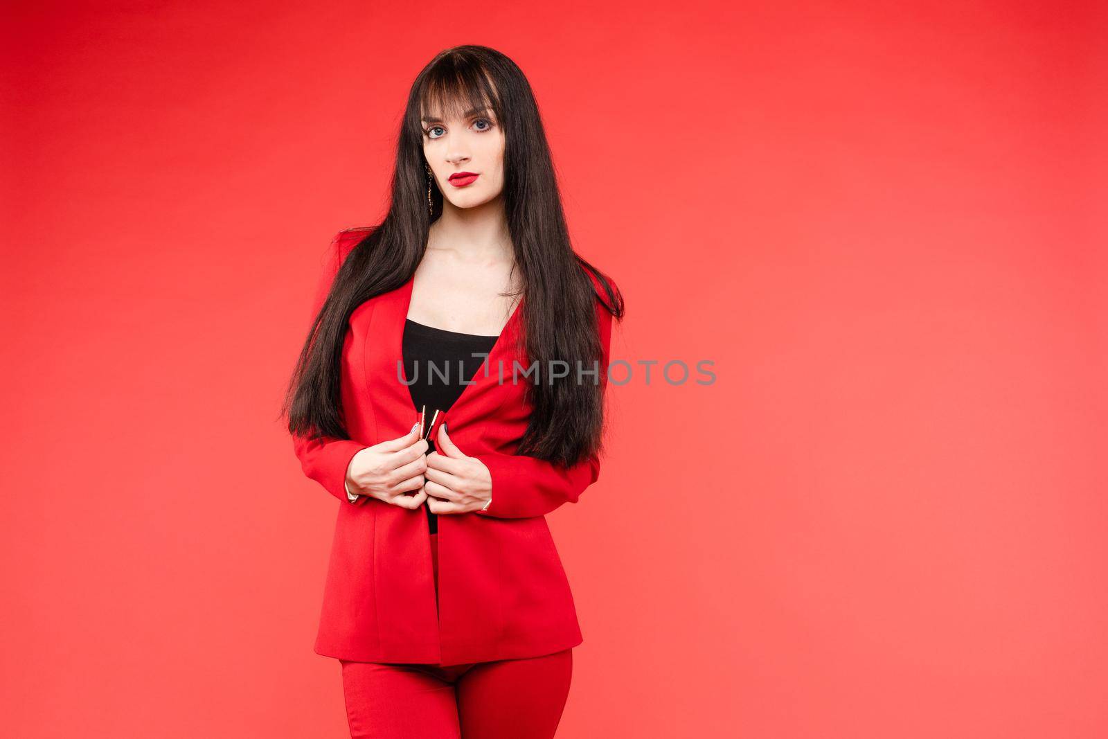 Young brunette model with blue eyes posing in fashionable clothes. Beautiful girl with long hair in elegant red costume looking at camera. Gorgeous lady standing near bunch of black and gold balloons.