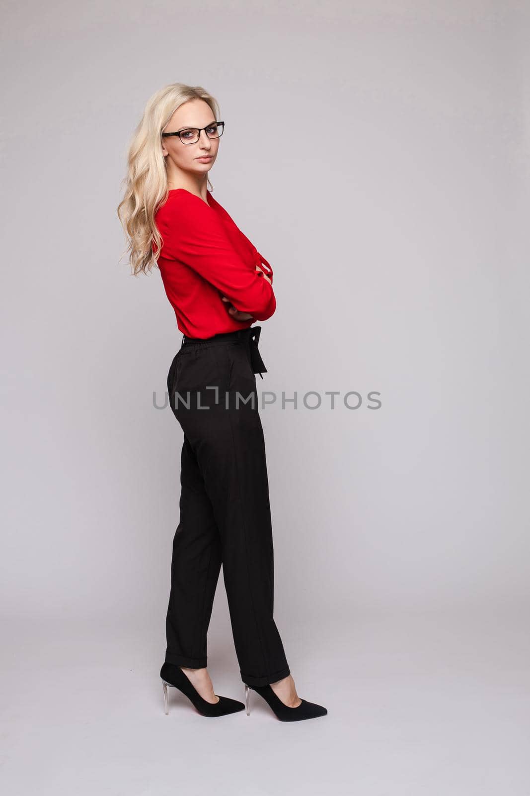 Front view of elegant successful female wearing smart outfit and glasses and posing on isolated background. Intelligent woman with folded arms looking at camera in studio. Concept of job and business.